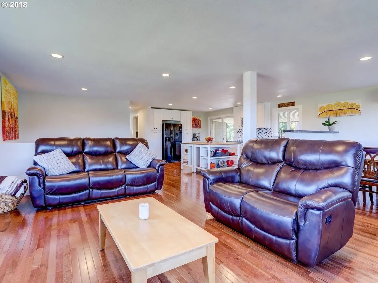Living room with hardwood floors, two brown leather sofas, a light wood coffee table, a colorful abstract painting on the wall, and a view into a kitchen area with white cabinets and a small island.