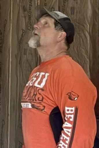 A man with a gray beard and mustache, wearing a black cap, an orange shirt with white and red text, and a jacket or sweatshirt tied around his waist, standing outdoors near a wooden surface.