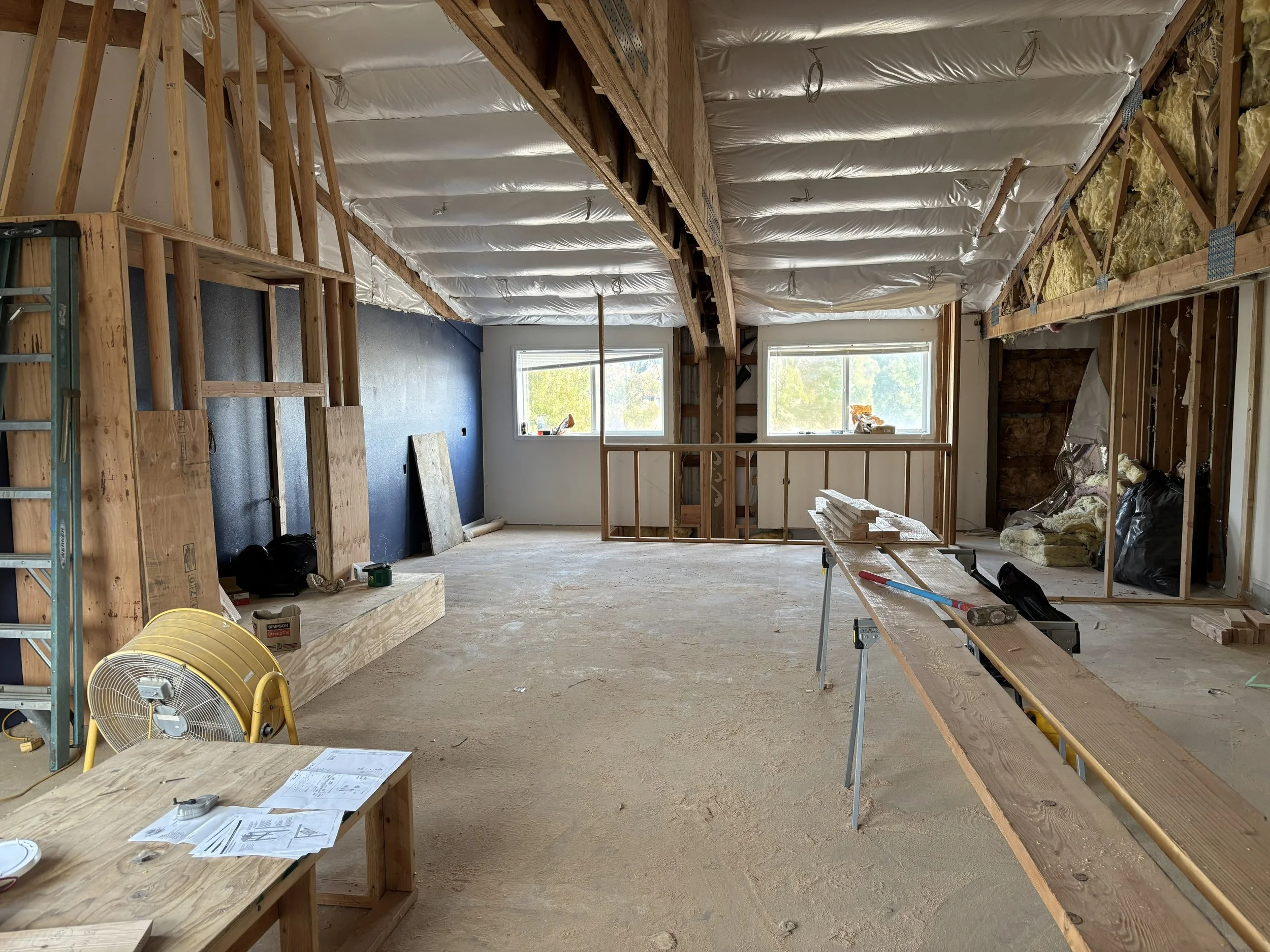 Interior view of a house under construction, showing exposed wooden framing, insulation, and partially finished walls. There are two windows at the back letting in natural light, with construction tools and materials scattered around.