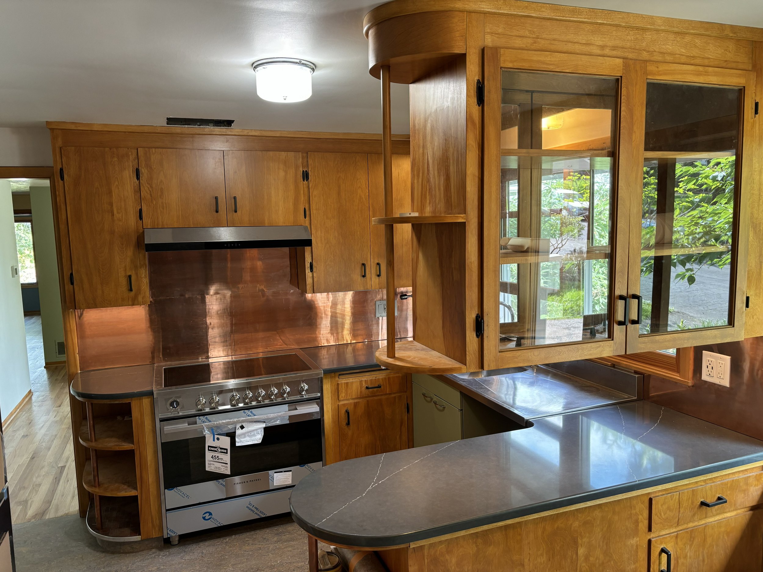 The image depicts a kitchen area with wooden cabinets, a stainless steel stove, and a curved bar counter with a dark marble countertop. There are large windows with wooden frames allowing natural light to illuminate the space, and some greenery visible outside.