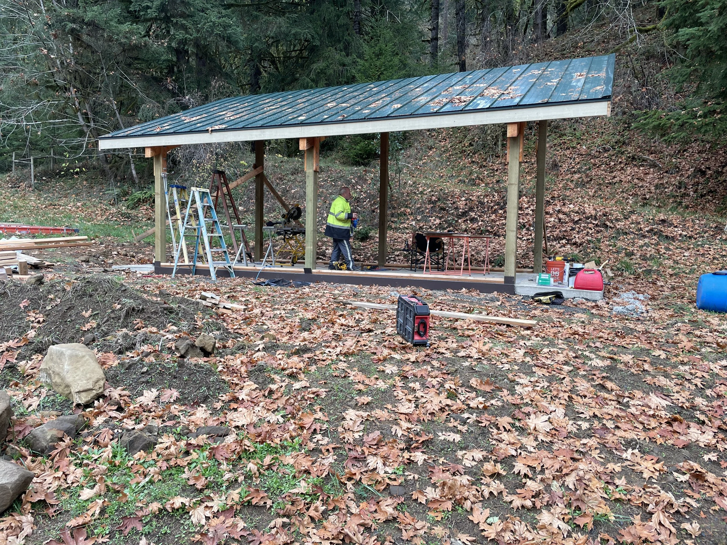 Construction workers building a shelter or small structure in a forested area with fallen leaves on the ground and trees in the background.