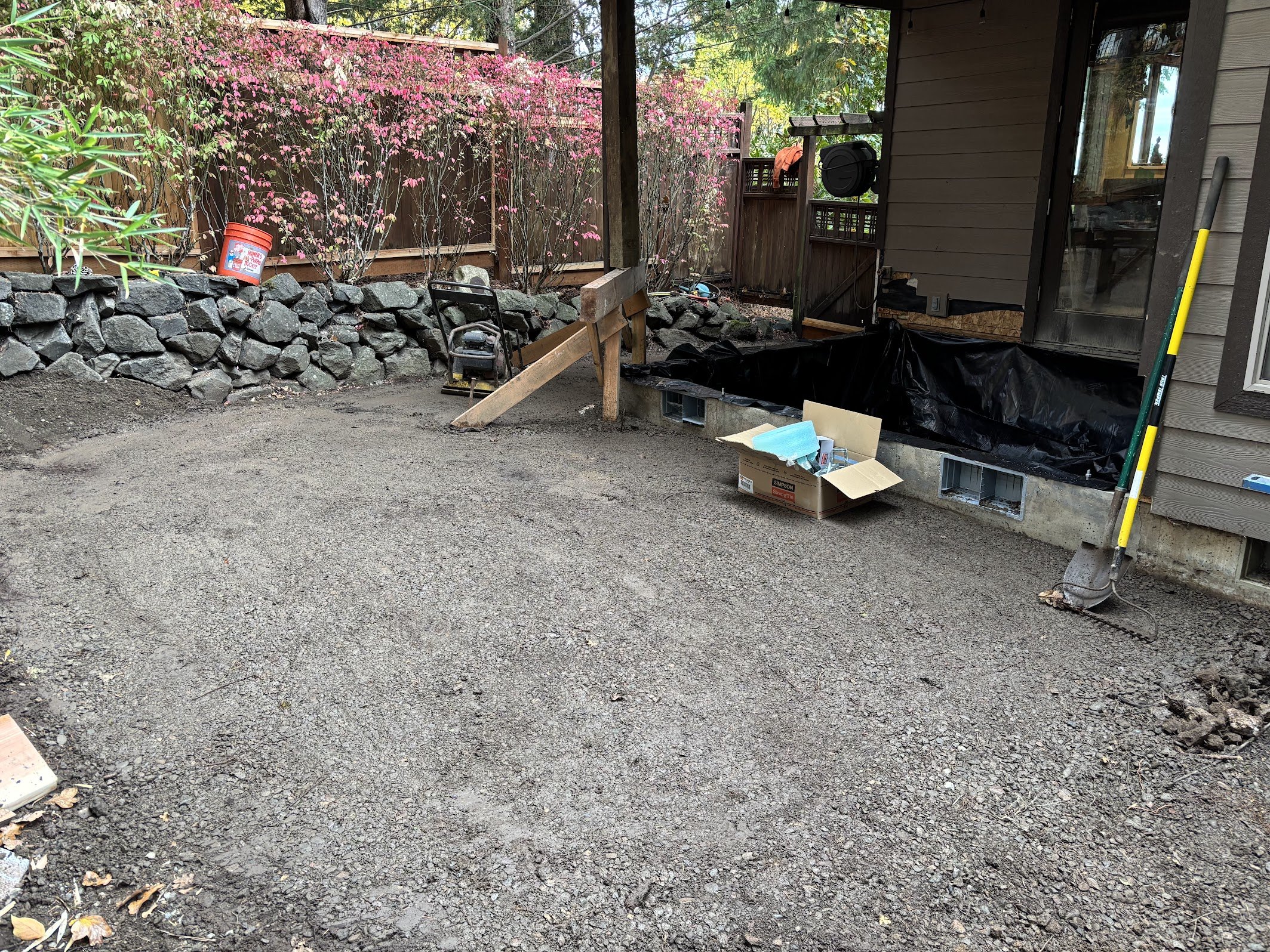 A backyard scene showing a wooden fence with pink flowering shrubs, a stone retaining wall, a partially dug hole with construction tools, a cardboard box, and a house exterior with siding and a door.