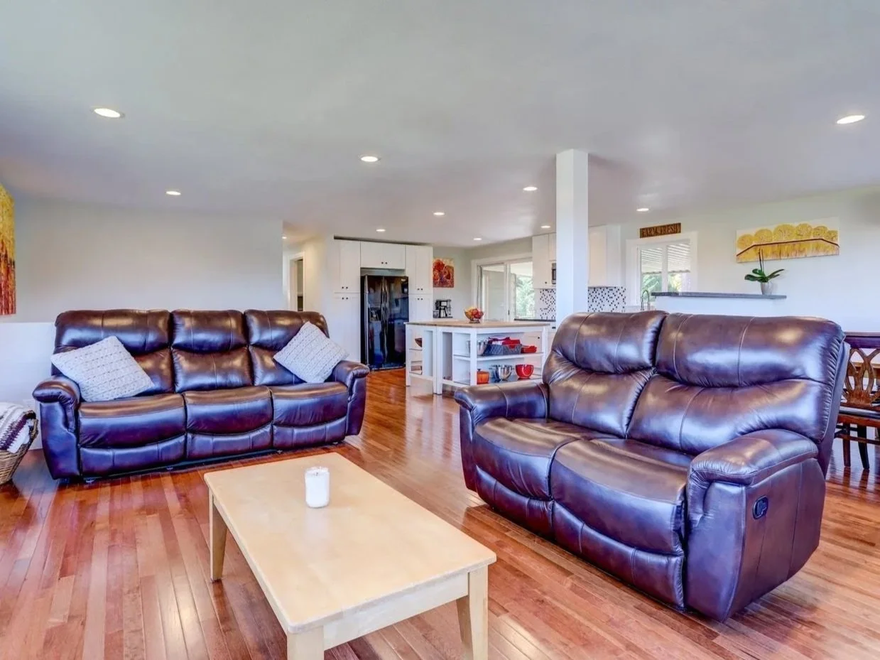 Living room with two brown leather sofas, a wooden coffee table, hardwood floors, and a view of an open kitchen with white cabinets, a refrigerator, and decorative wall art.