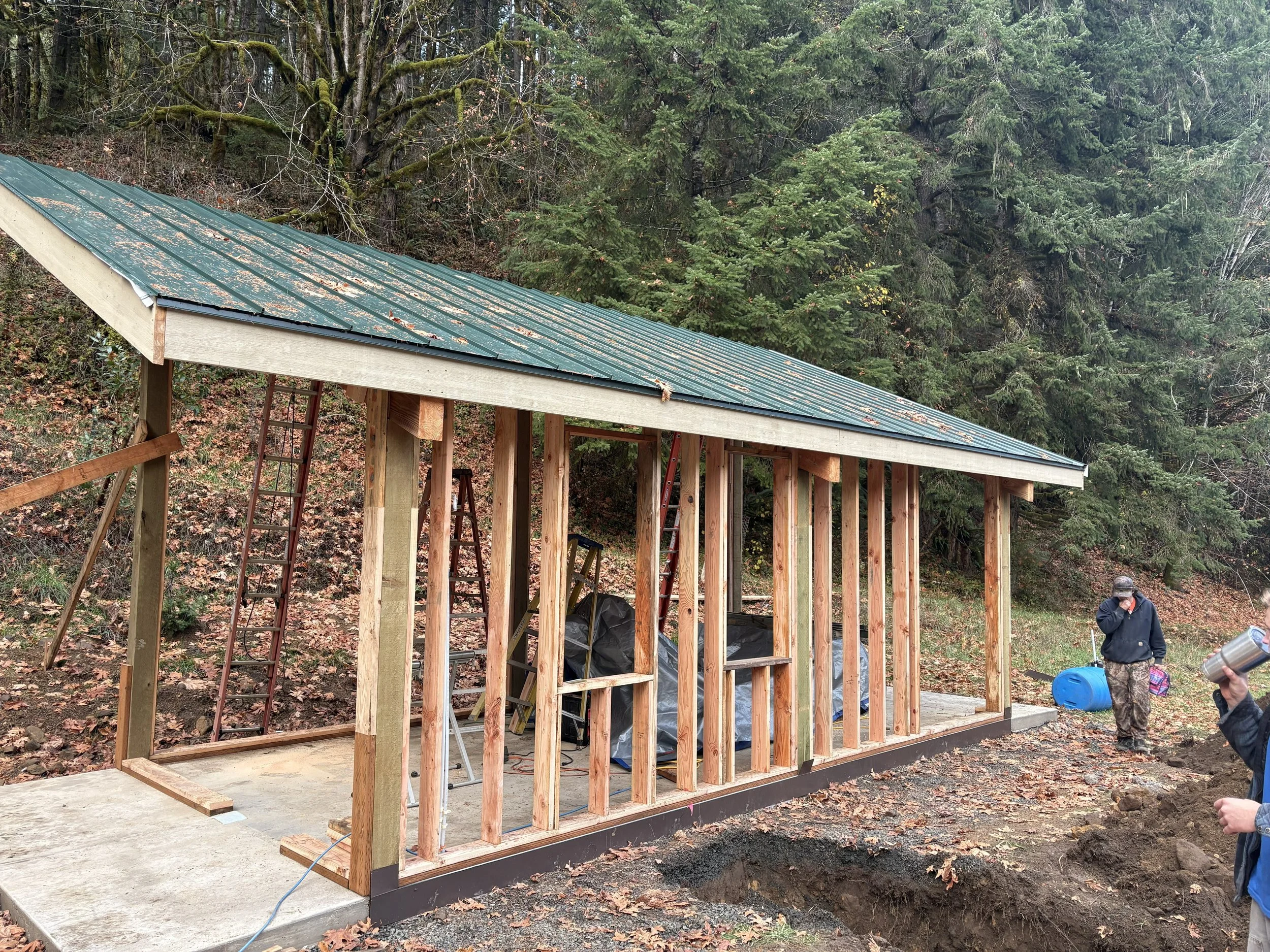 Wooden structure under construction with a metal roof, surrounded by trees, and construction workers nearby.