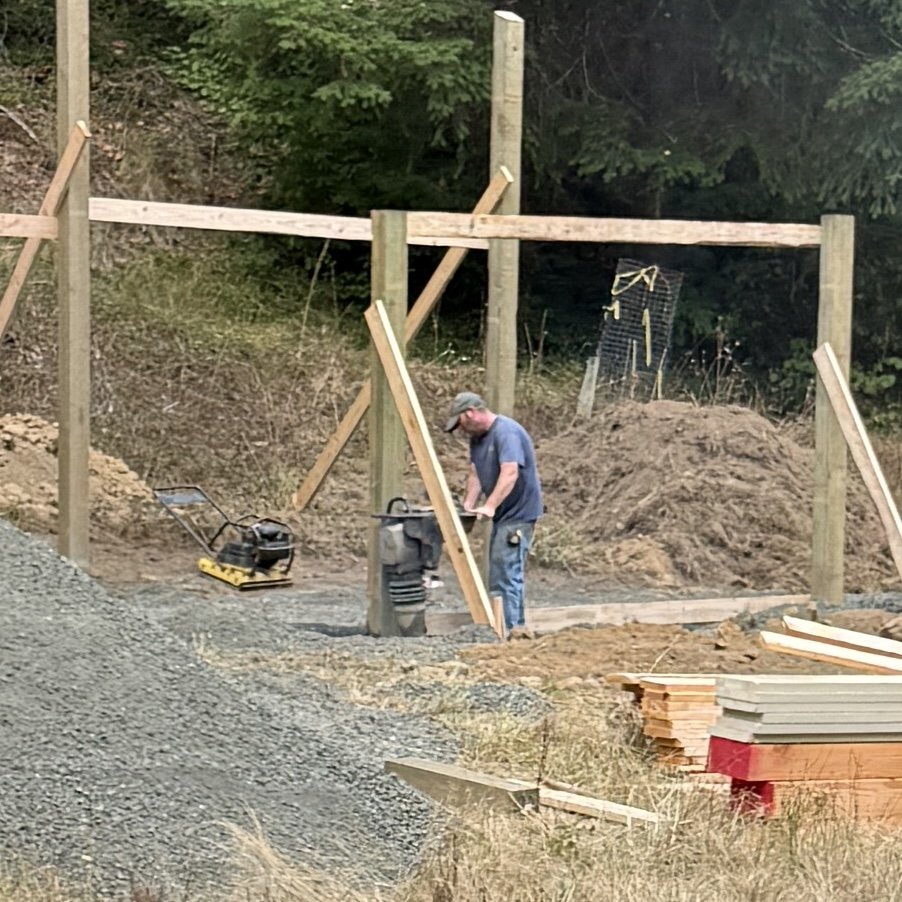 A construction worker working on a wooden fence structure outdoors, with trees in the background and construction materials and tools on the ground.