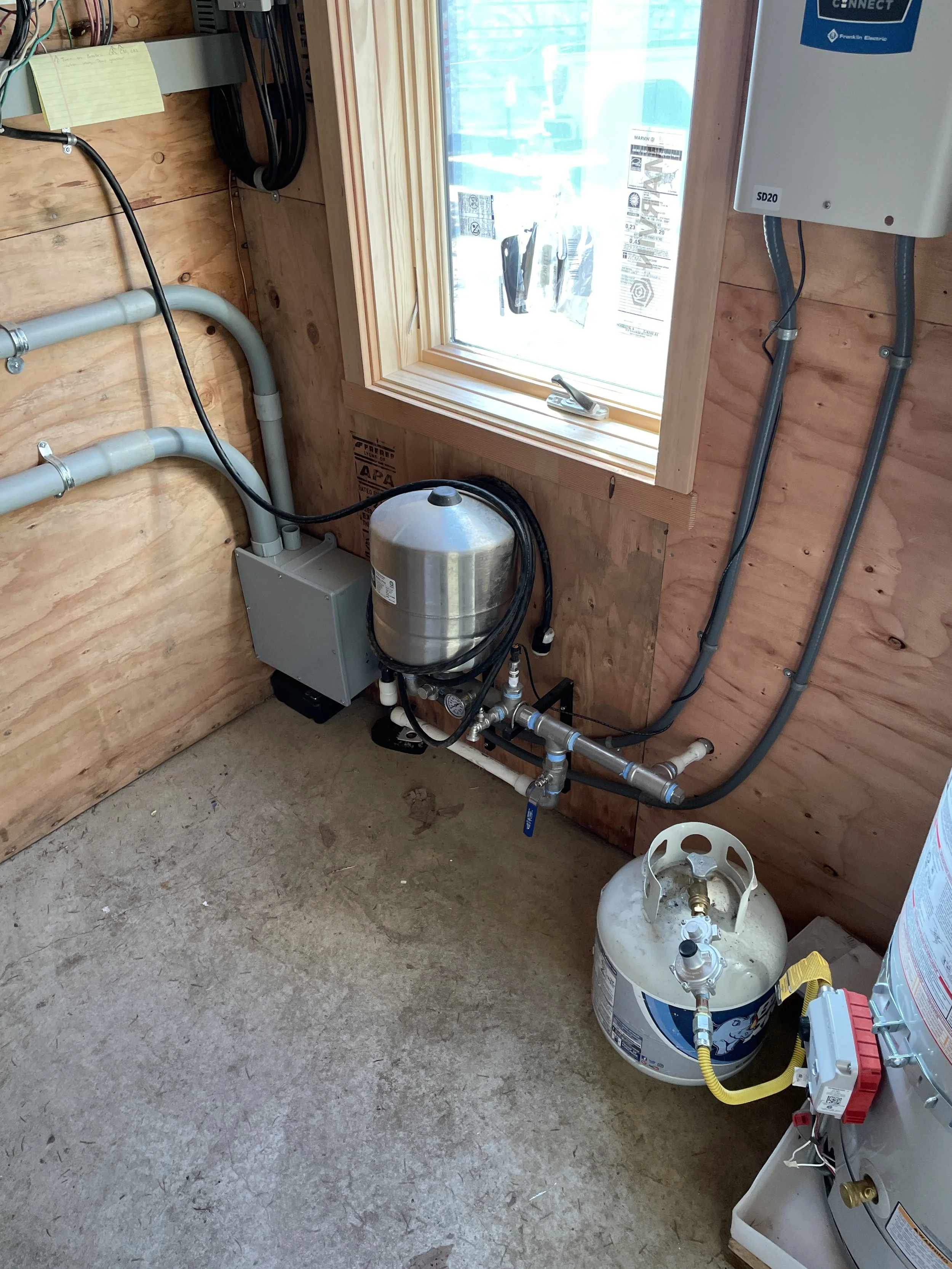 A utility room with exposed wooden walls and a concrete floor. It contains plumbing and electrical wiring, a small window, and a propane tank on the floor connected by yellow and gray hoses to a heater or boiler.