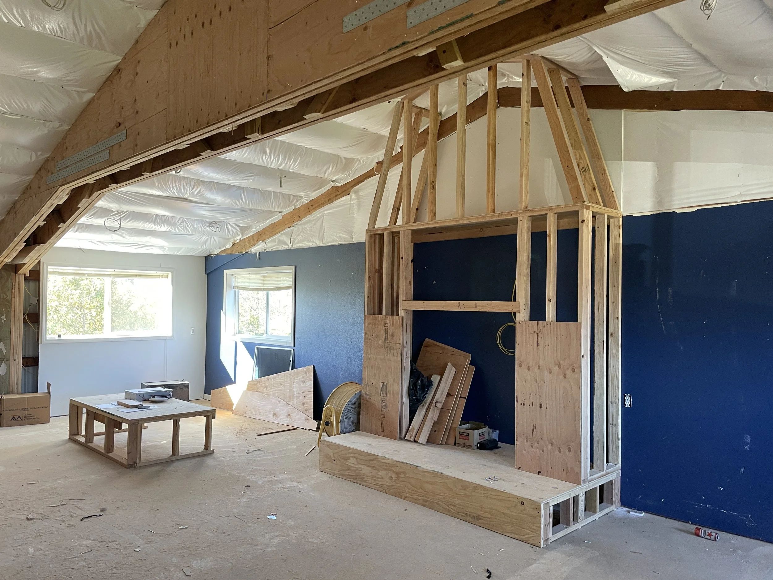 Interior view of a house under construction, featuring wooden framing for a fireplace, stairs, and a window with sunlight coming through.