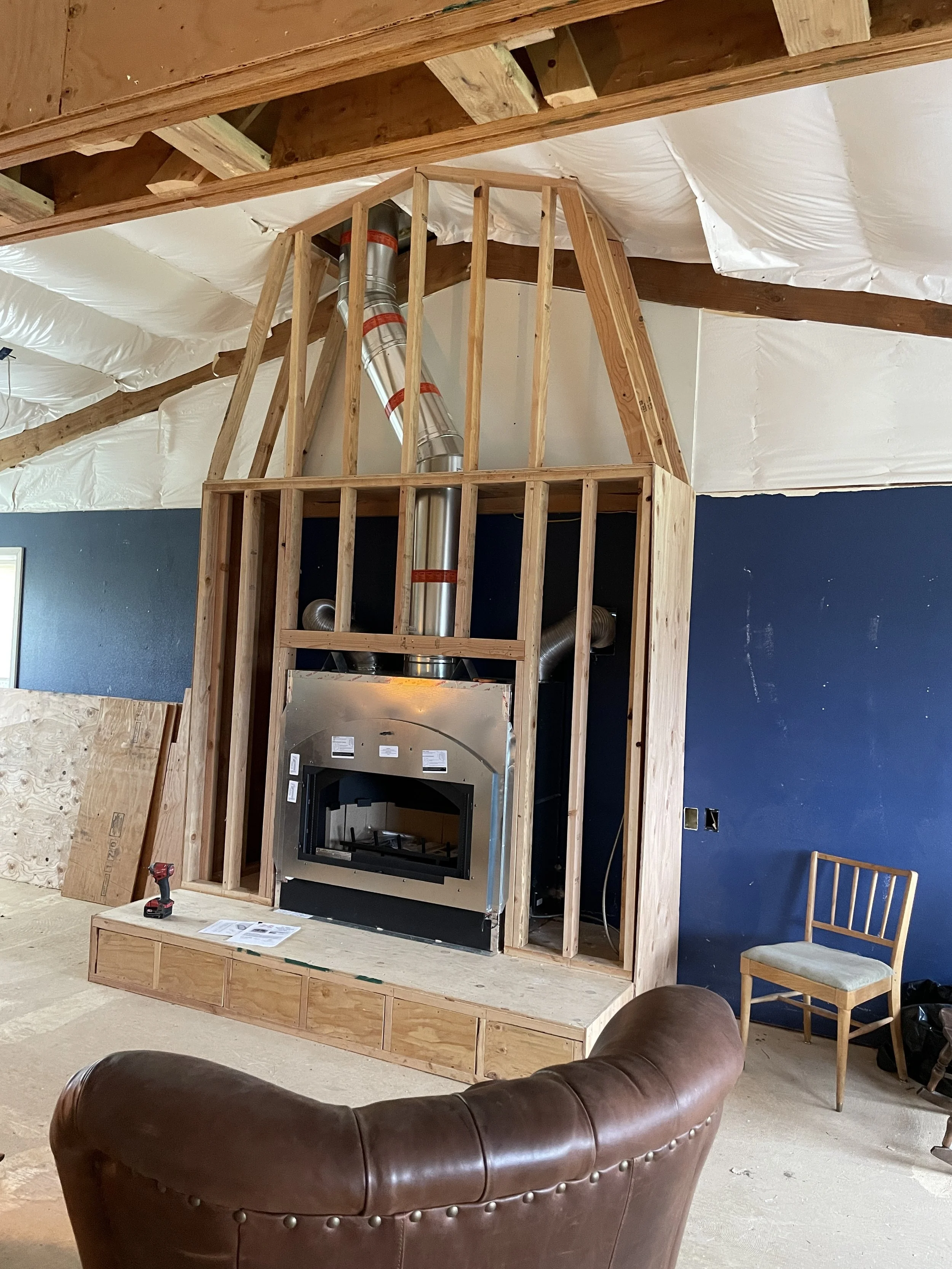 Construction of a framed wall with a fireplace and ductwork in a home under renovation, with a brown leather couch and a wooden chair in the foreground.