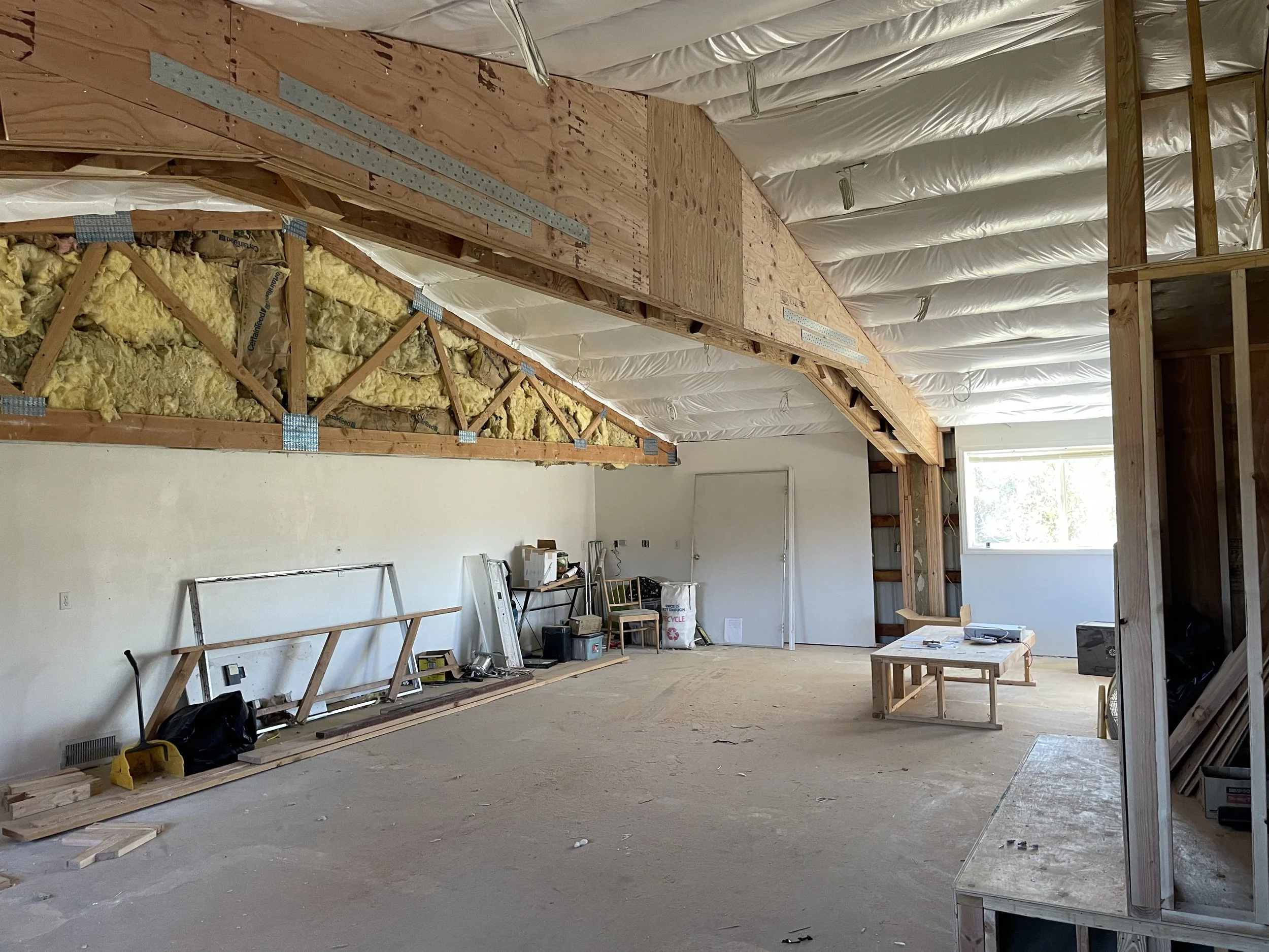 Interior of a room under construction with exposed wooden framing, insulation, and partially finished ceiling. Construction tools and materials are scattered on the floor.