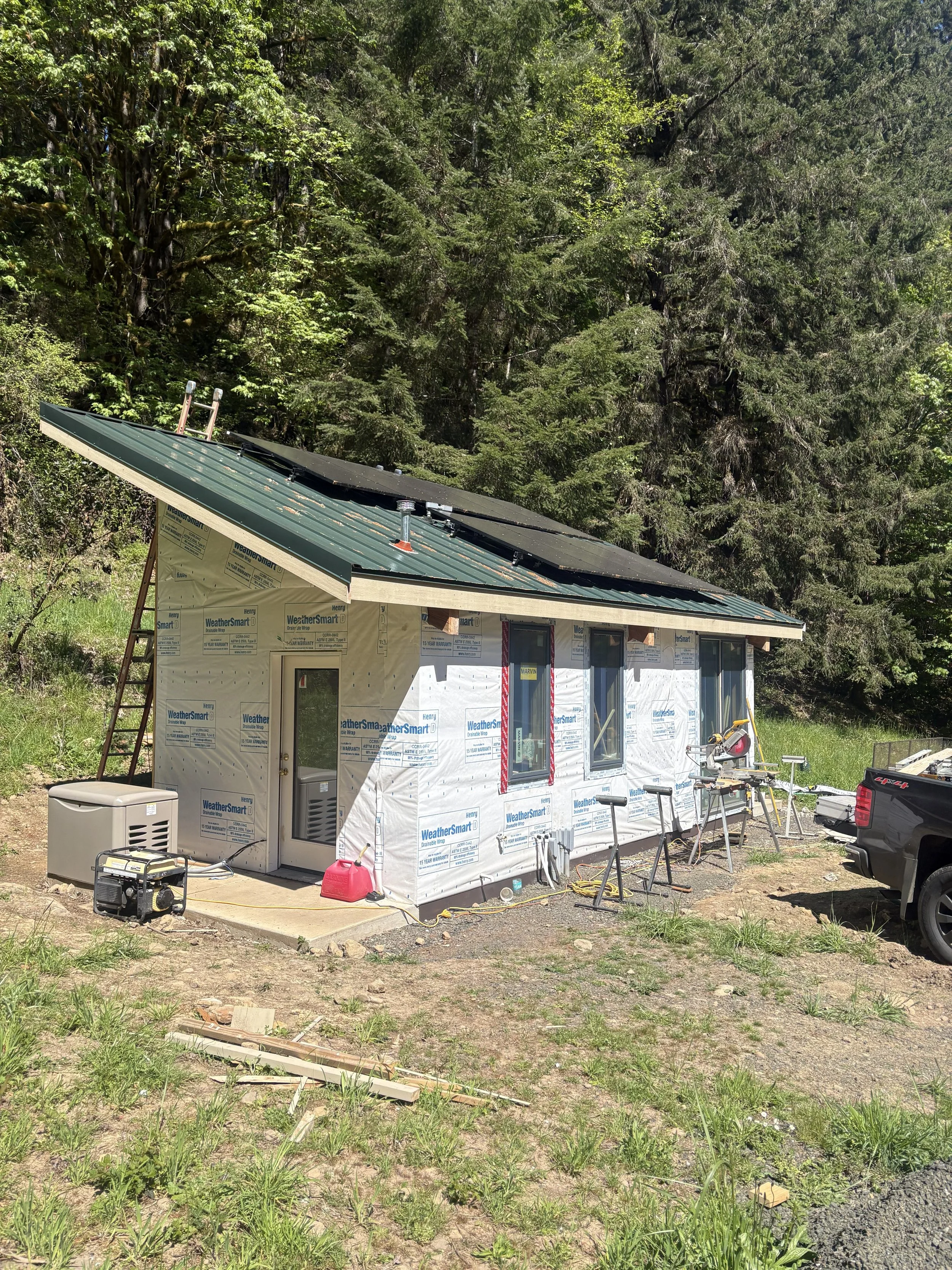 Construction of a house with a green metal roof with solar panels in a rural area, surrounded by trees.