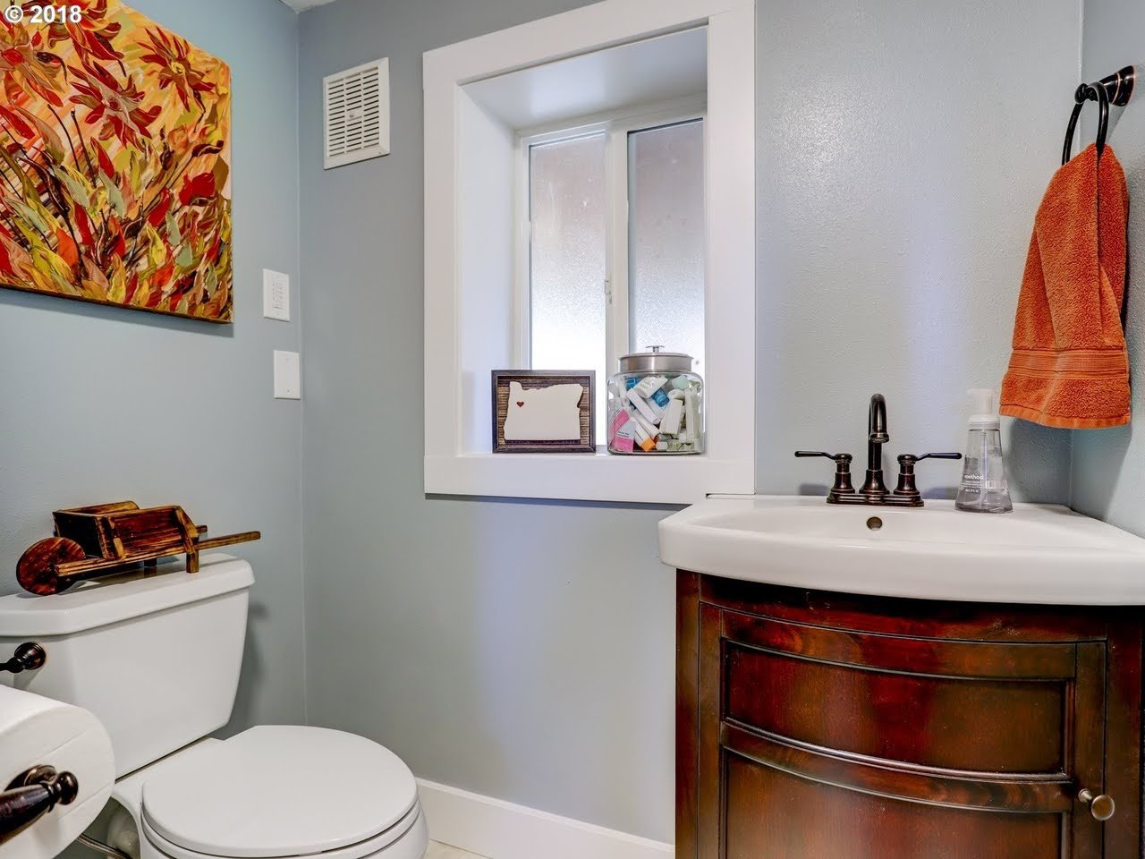 Small bathroom with light blue walls, a white toilet, a dark wood vanity with a white sink, and a small window above the sink. Decor includes a colorful wall art, an orange towel, and decorative items on the window ledge.