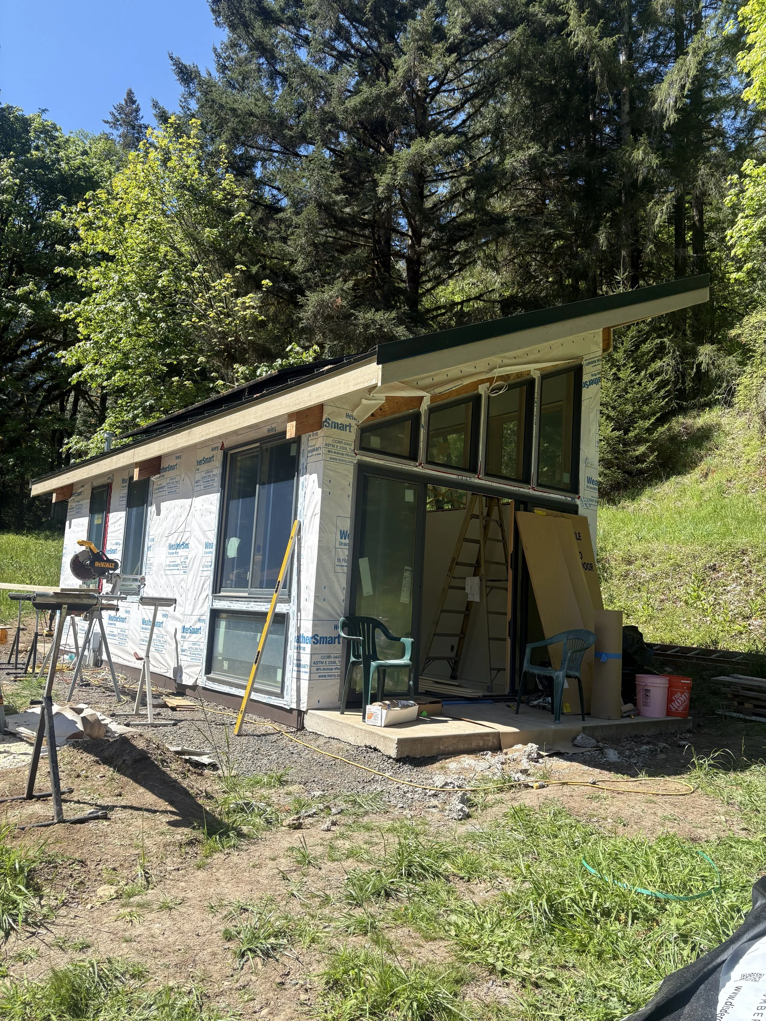 Construction site of a house with large windows and a sloped roof, surrounded by trees, construction tools, and materials.