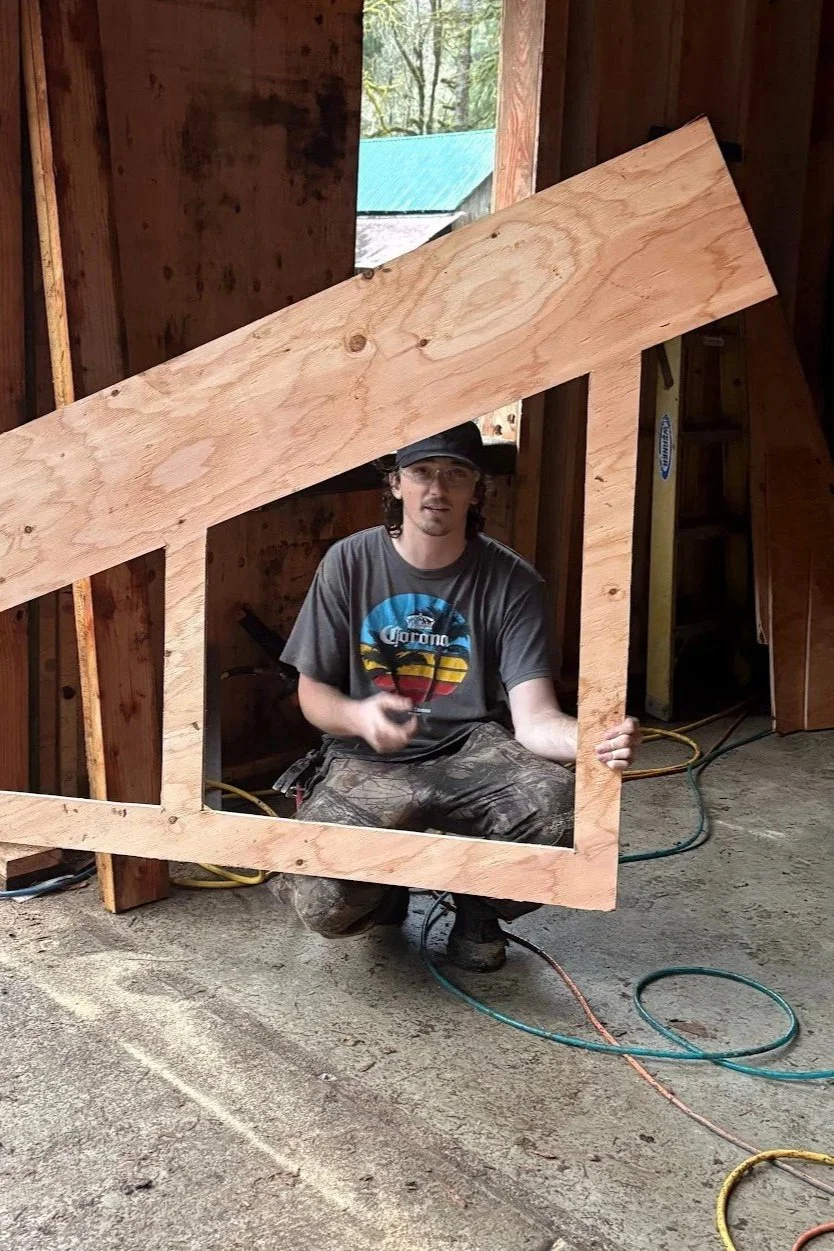 A man wearing a Corona t-shirt and camouflage pants is kneeling inside a wooden frame in a construction site, holding a piece of wood. The frame has a large cutout, and part of the unfinished wooden wall and a window with a view of trees and a green roof are visible.