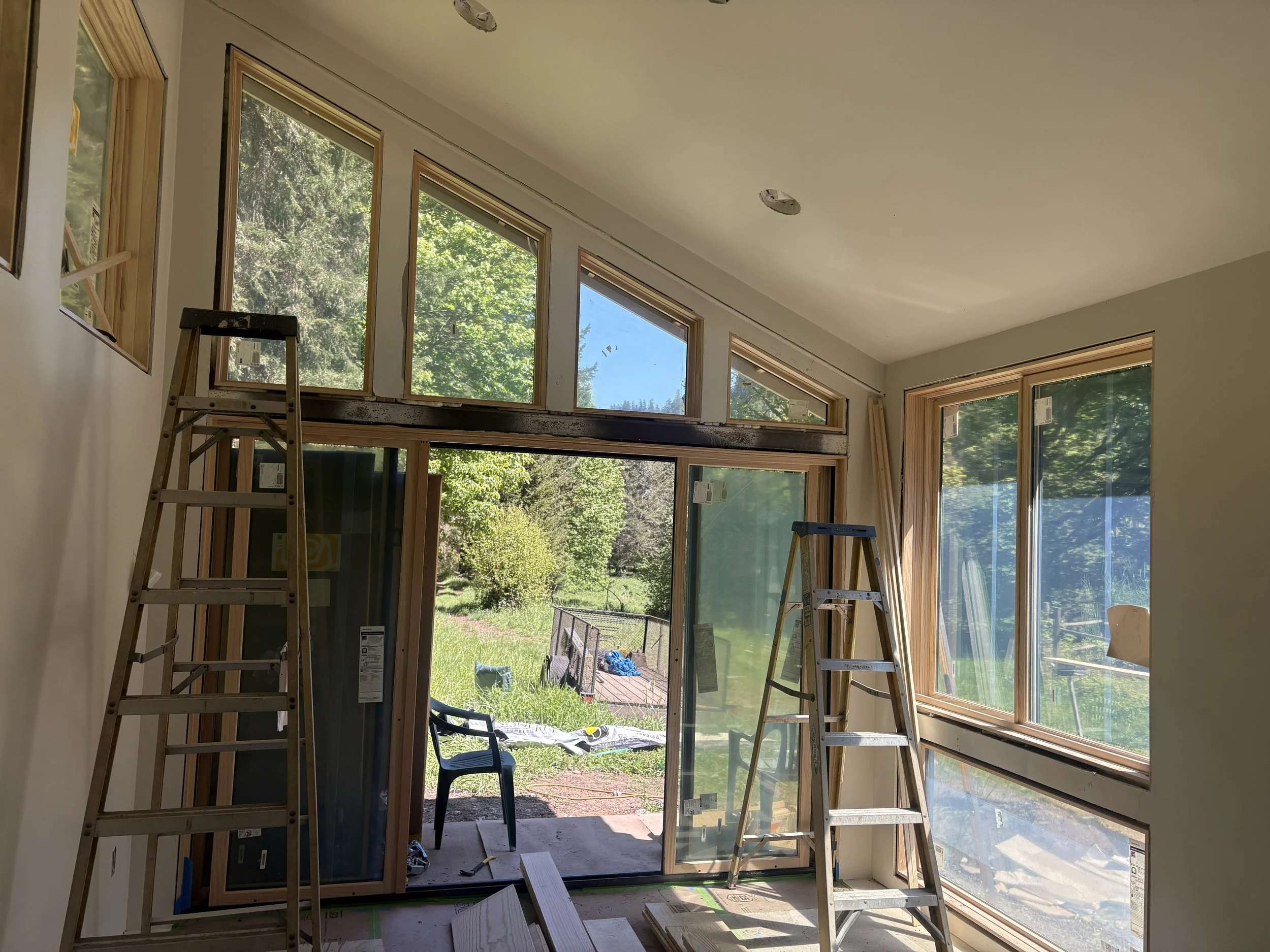 Interior of a house under construction with large windows, ladders, and construction materials, overlooking a green yard outside.