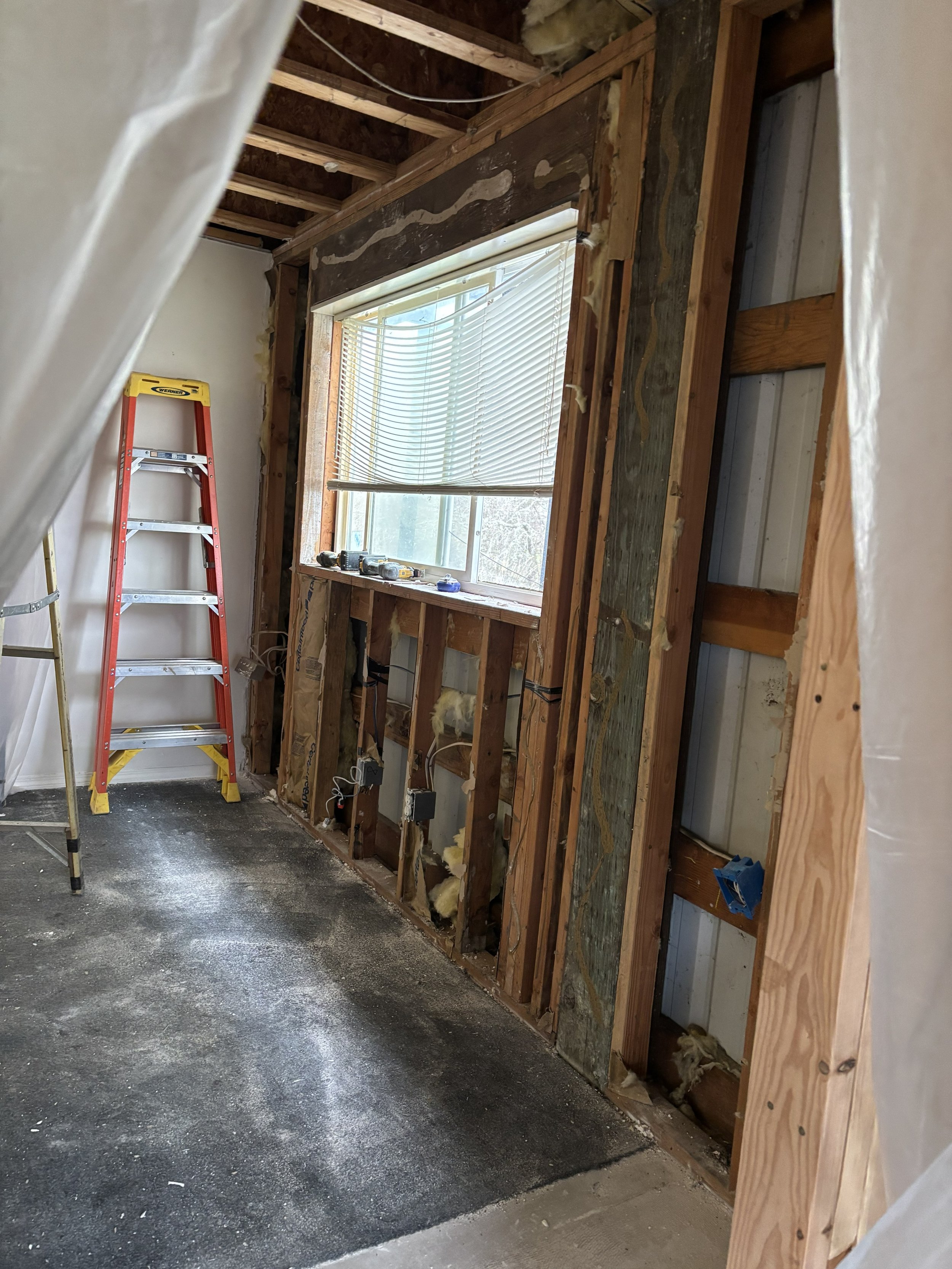 Interior of a room under renovation with exposed wooden framing, a window with blinds, a ladder, and construction tools.