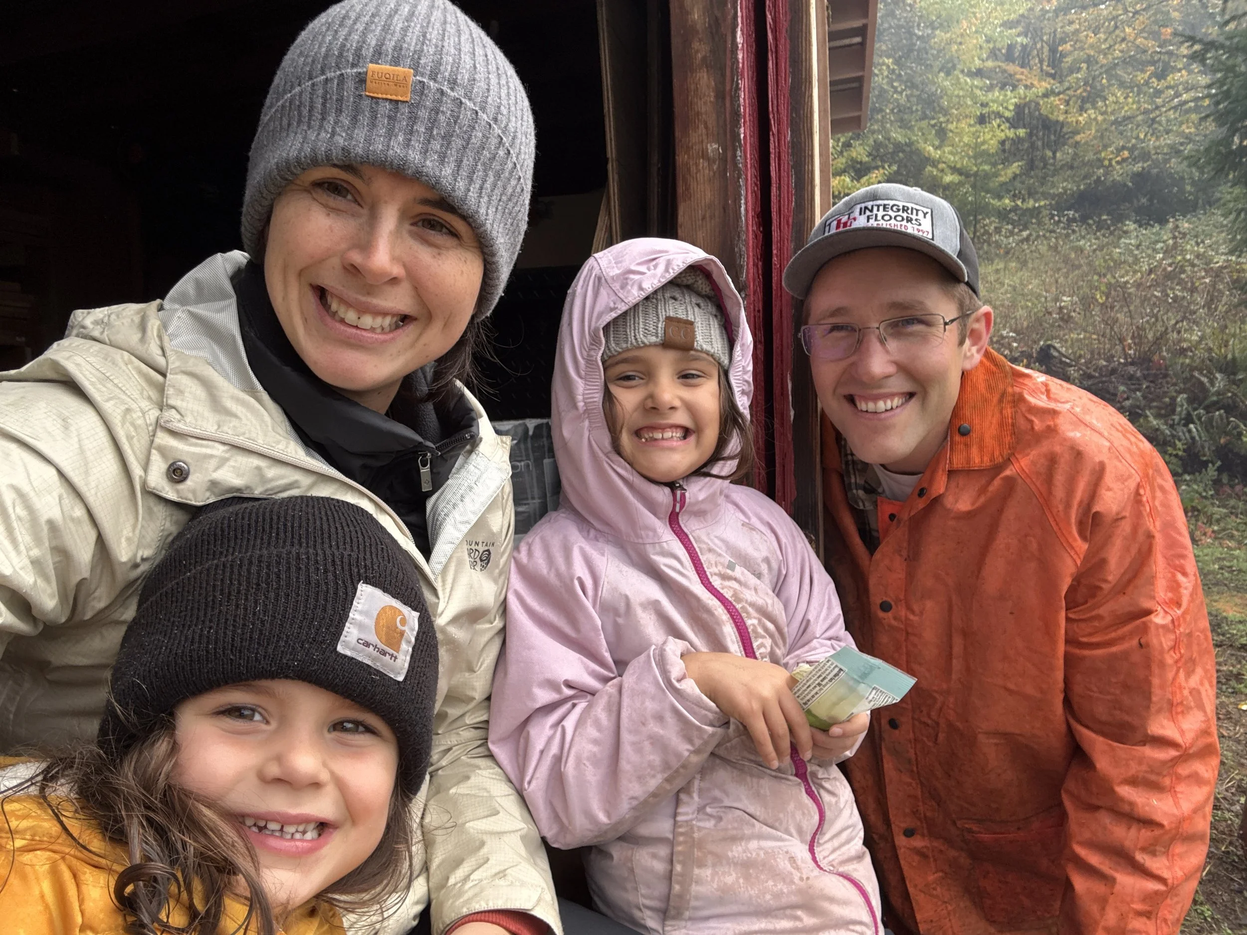 A smiling family of four, including two young girls and two adults, standing outdoors on a cloudy day with autumn trees in the background. They are dressed warmly, with beanies and jackets.