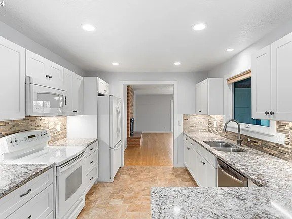 Kitchen with white cabinets, granite countertops, tiled backsplash, stainless steel sink, and white appliances, including refrigerator, microwave, and stove. Hardwood floor in the background leading to another room.