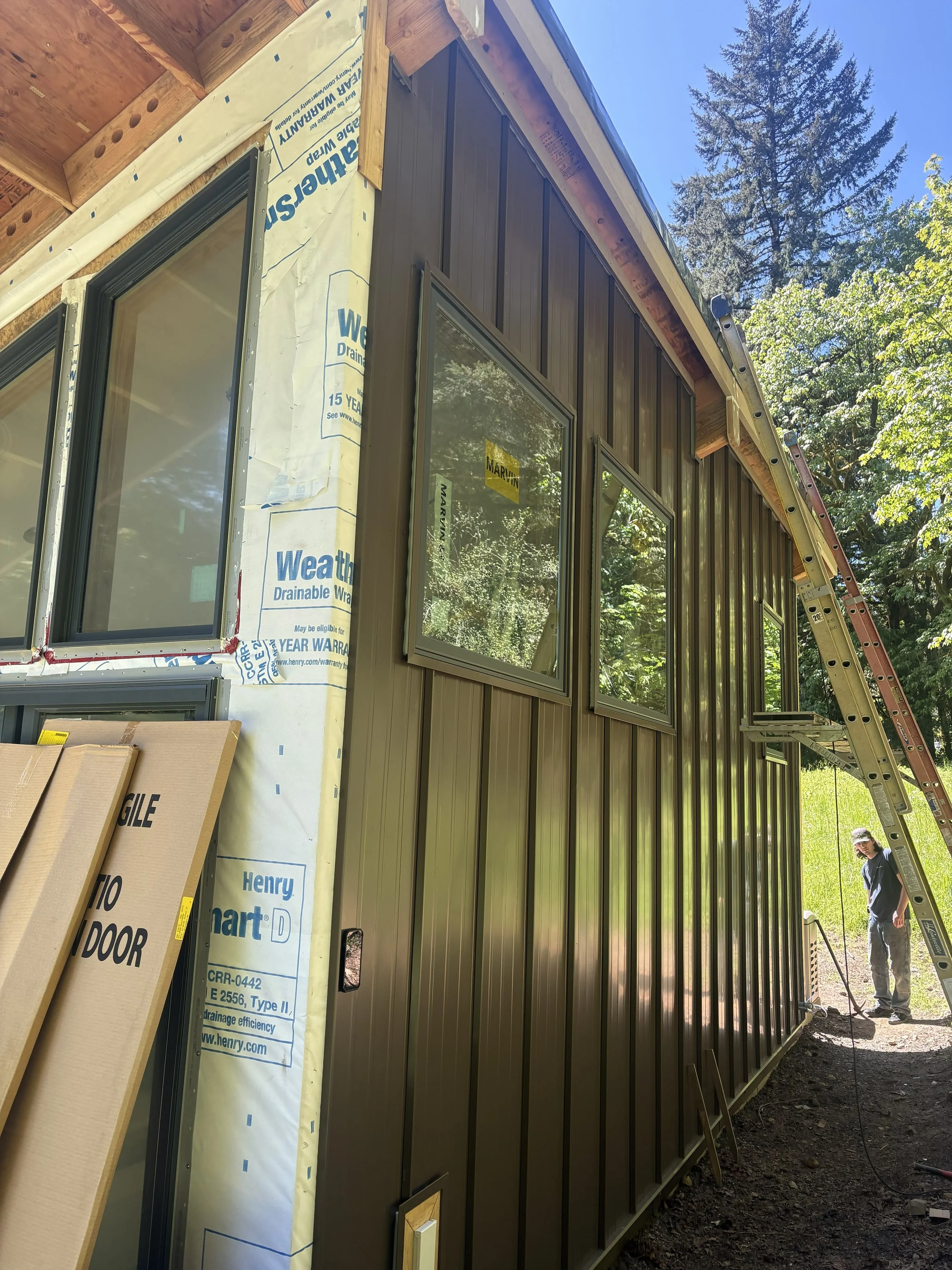 Side view of a house under construction with dark brown metal siding and window frames, building materials and a ladder visible, and a worker standing outside with a power tool, trees and blue sky in the background.