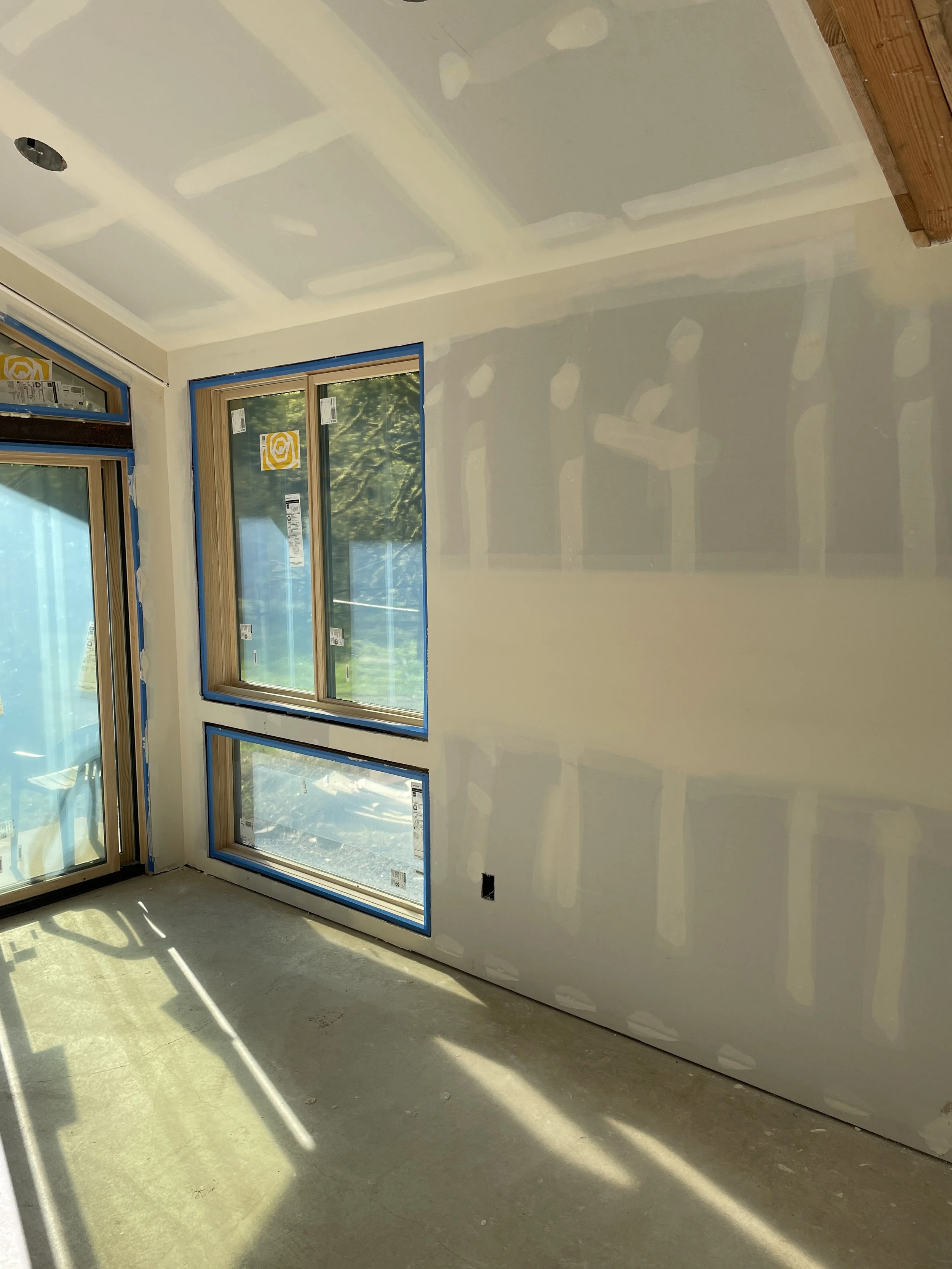 Interior room under construction with unfinished drywall, new windows with blue tape around the edges, and sunlight casting shadows on the concrete floor.
