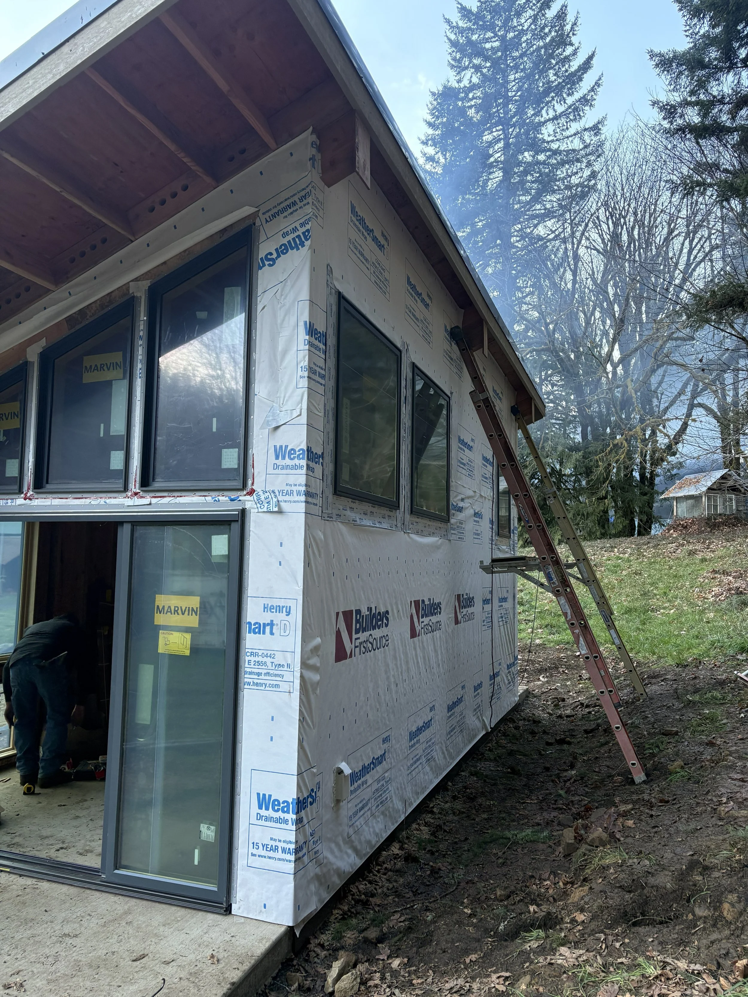 Construction site of a house with weather barrier wrap on the exterior walls, a sliding glass door installed, and a leaning ladder against the house. Trees and a shed are visible in the background.