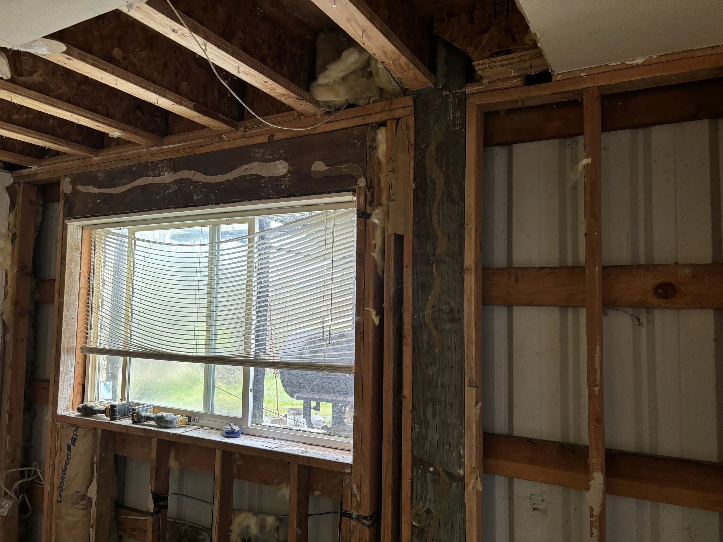 The interior of a room under renovation with exposed wooden framing, insulation, and a window with blinds. Construction tools are placed on the window sill.