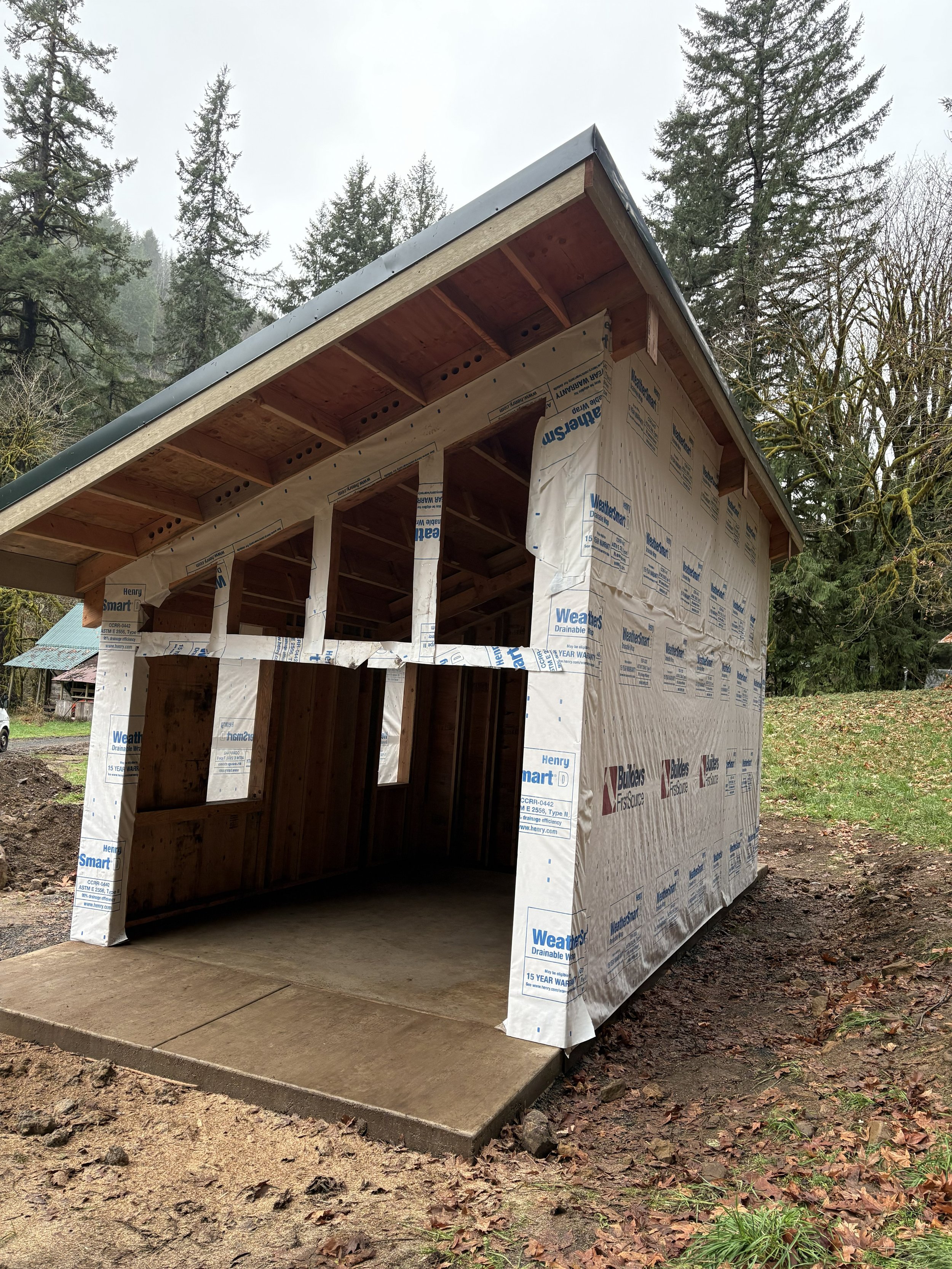 A small wooden structure under construction with weather-resistant wrap on the exterior, a concrete slab foundation, and a pitched roof amid trees in a wooded area.