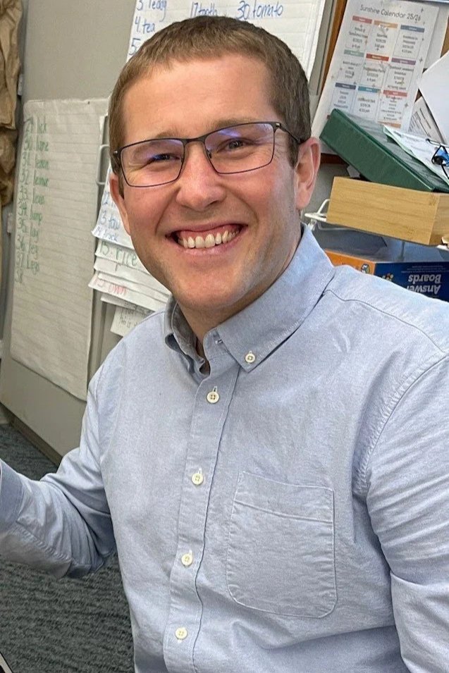 Smiling man with glasses sitting at a desk in a classroom or office setting, with charts and papers on the wall behind him.