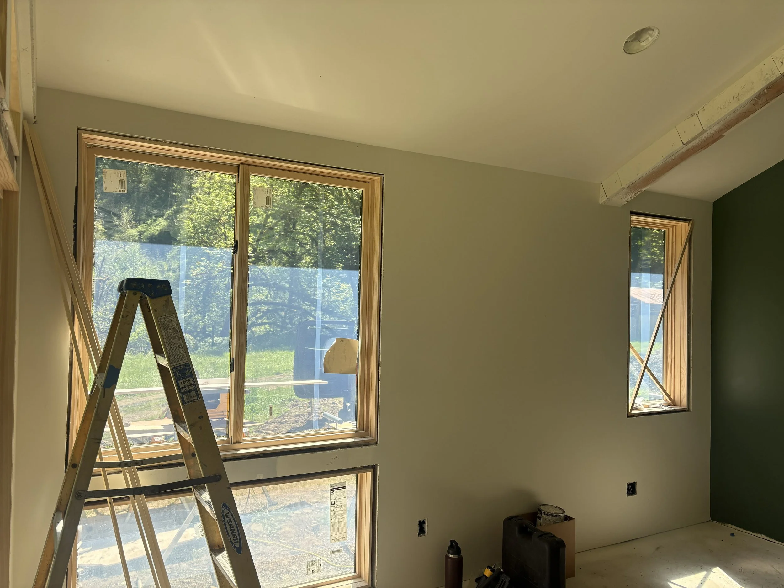Interior of a room under construction with large windows and a ceiling beam. Construction tools and materials are visible on the floor.