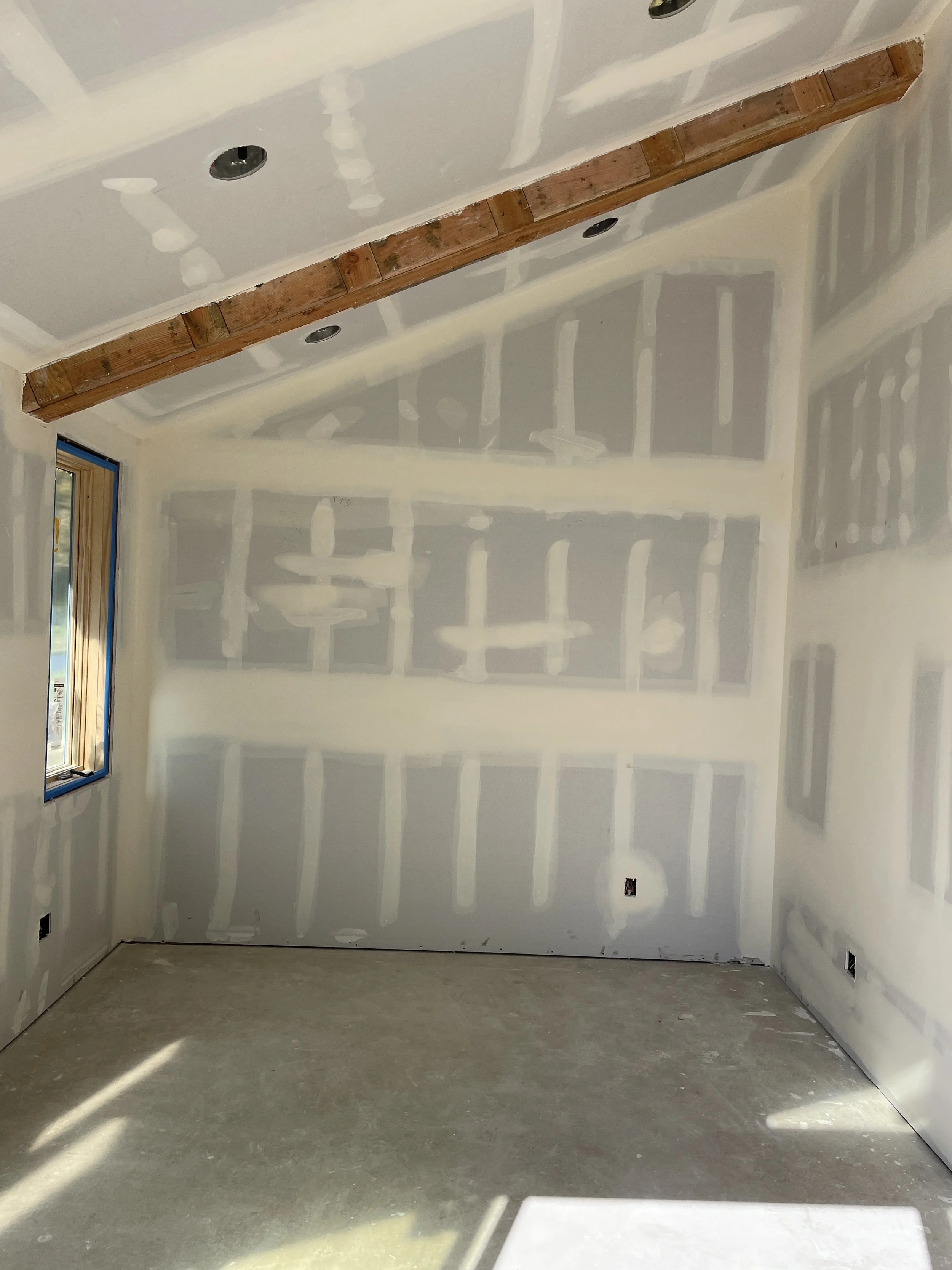Empty room under construction with drywall walls that have been taped and mudded, a single window with natural light, an exposed ceiling beam, no finished flooring, and electrical outlets installed.