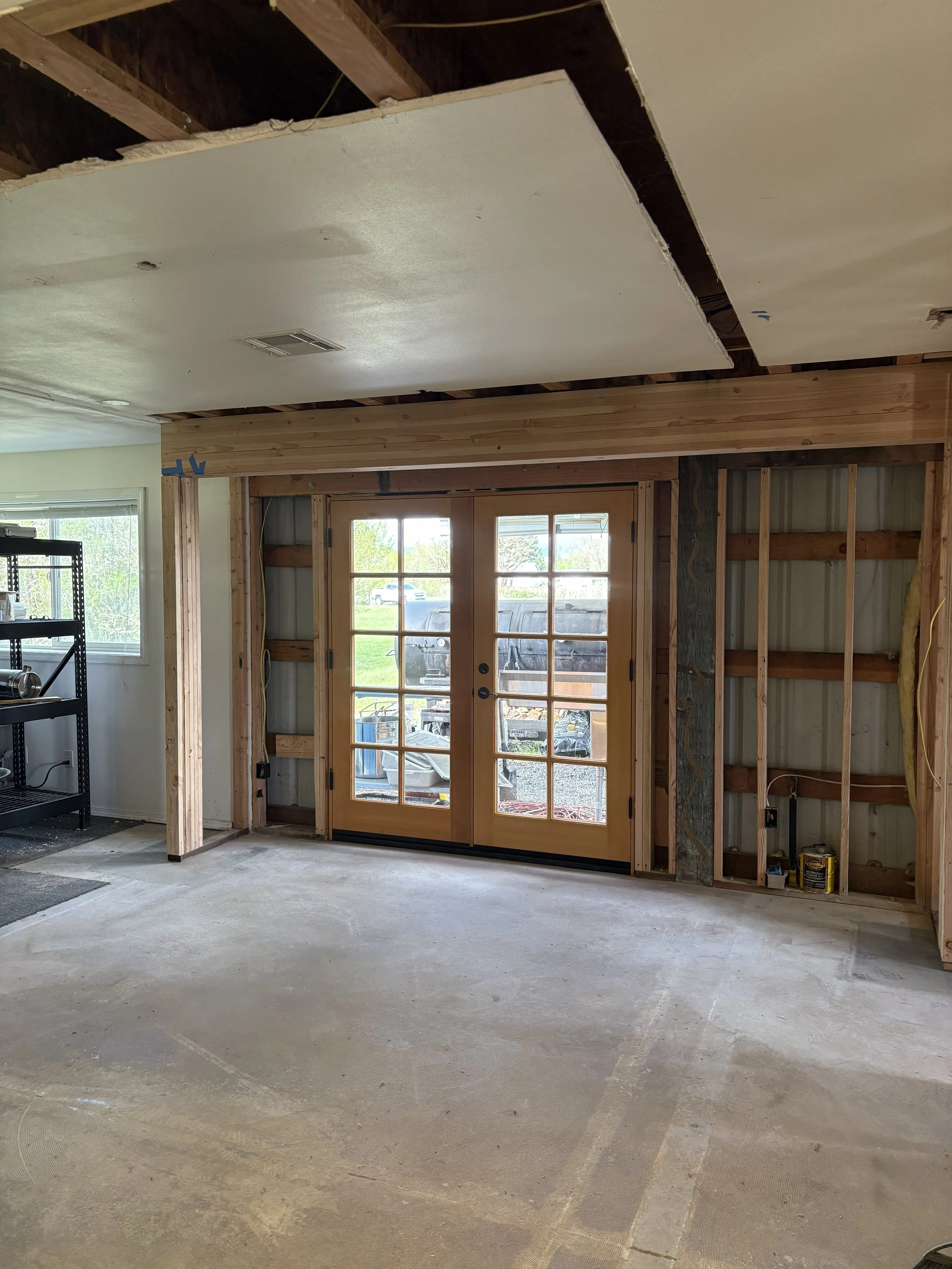Interior of a room under construction with exposed wooden framing, partial drywall, a double glass door, and an unfinished ceiling.