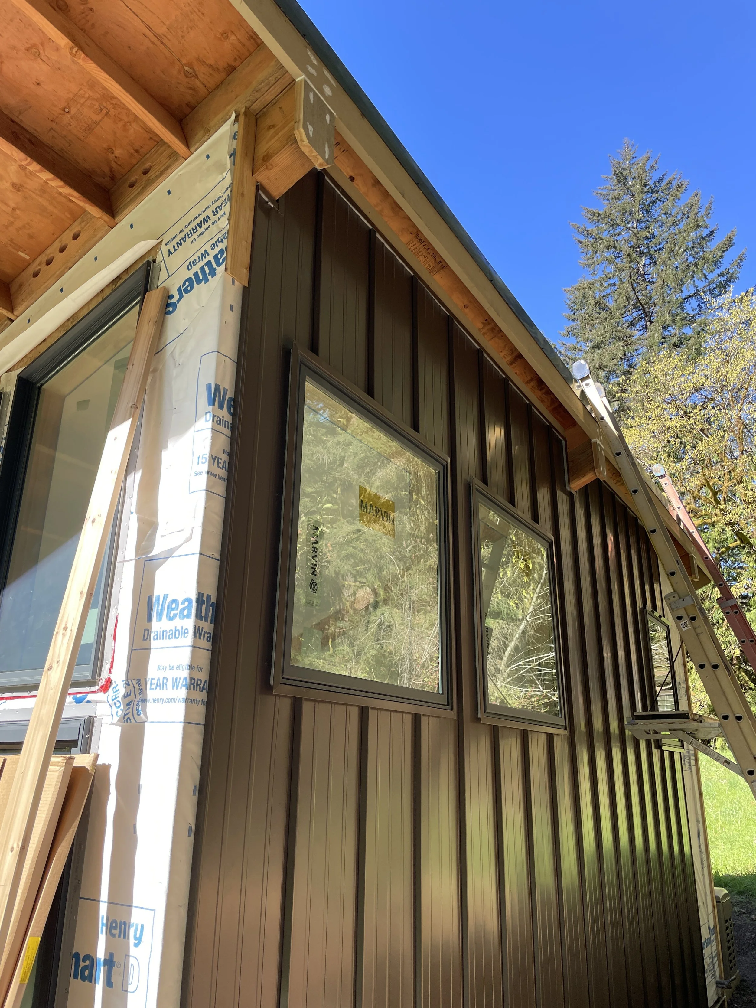 A house under construction with partially installed siding, windows, and an overhead roof. The exterior wall shows a combination of weather-resistant house wrap and metal siding, with a ladder leaning against the side. Trees and a clear blue sky are 