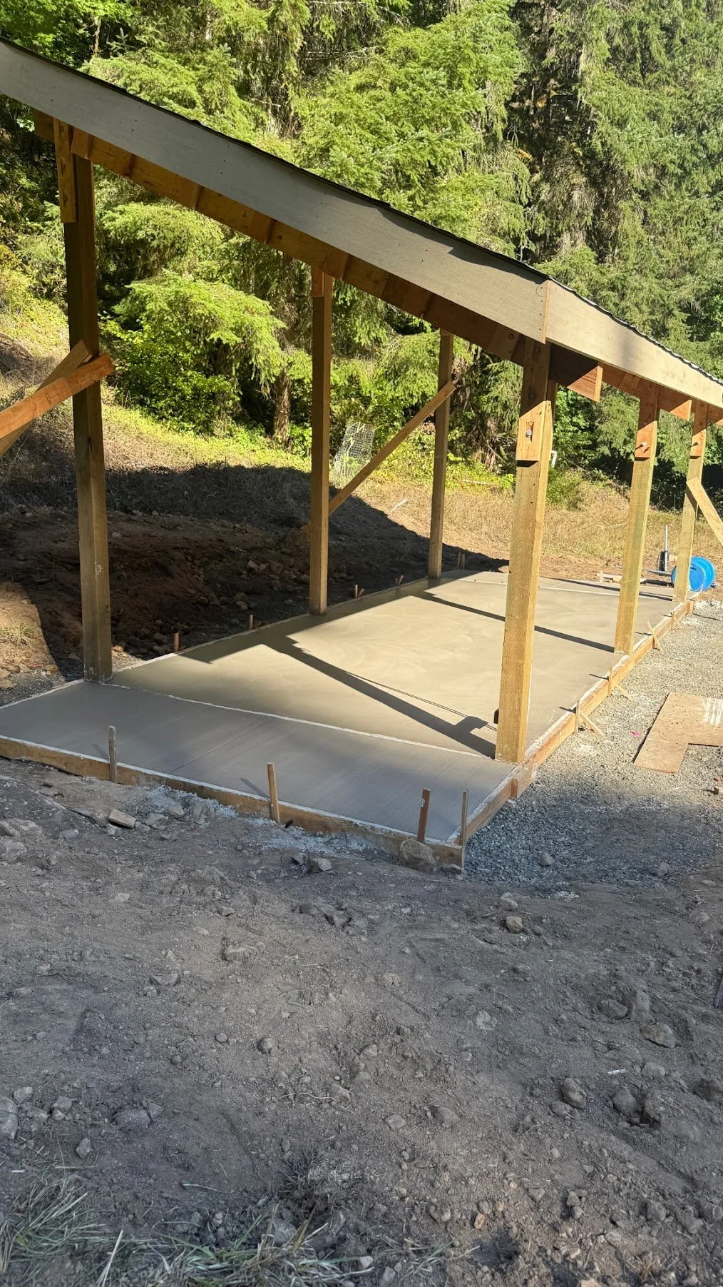 Construction site with a concrete slab foundation and wooden framing for a new building or shelter, surrounded by dirt and trees.