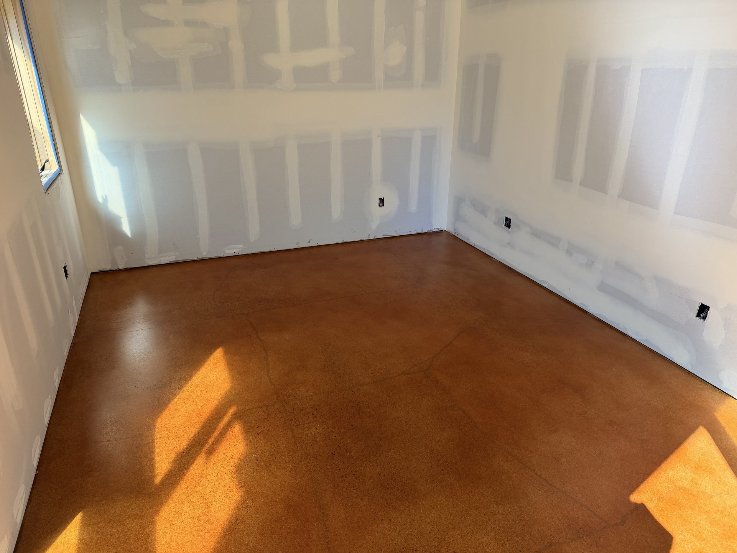 Empty room with a wooden floor, white walls with drywall seams, a small window on the left, and electrical outlets on the walls. Concrete slab has a copper, orange, rustic, polished was finish.