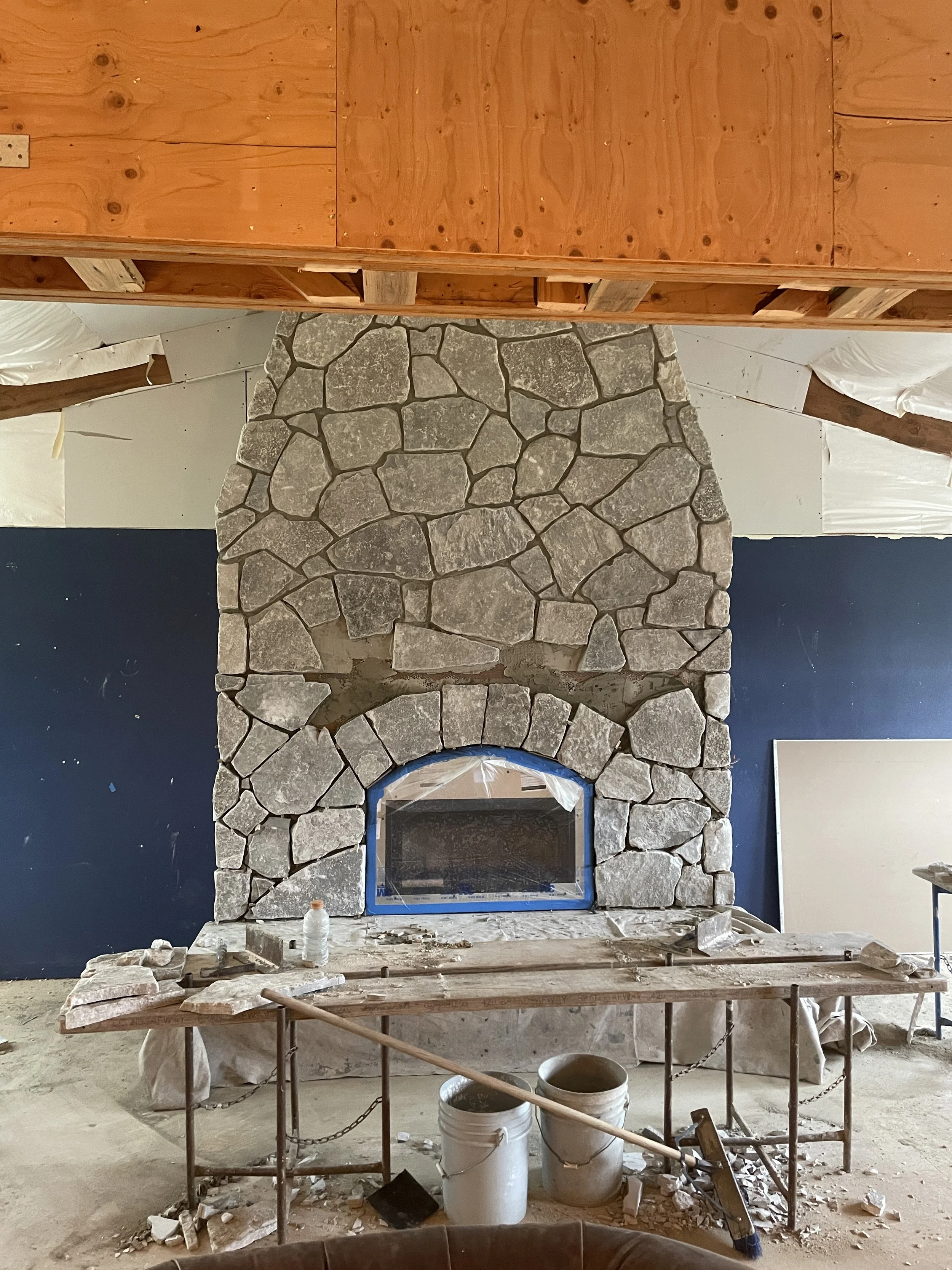 Under construction indoor space featuring a stone fireplace. The fireplace is made of irregularly shaped stones and has an arched opening. The area around the fireplace is under renovation, with construction tools, buckets, and debris on the floor. A