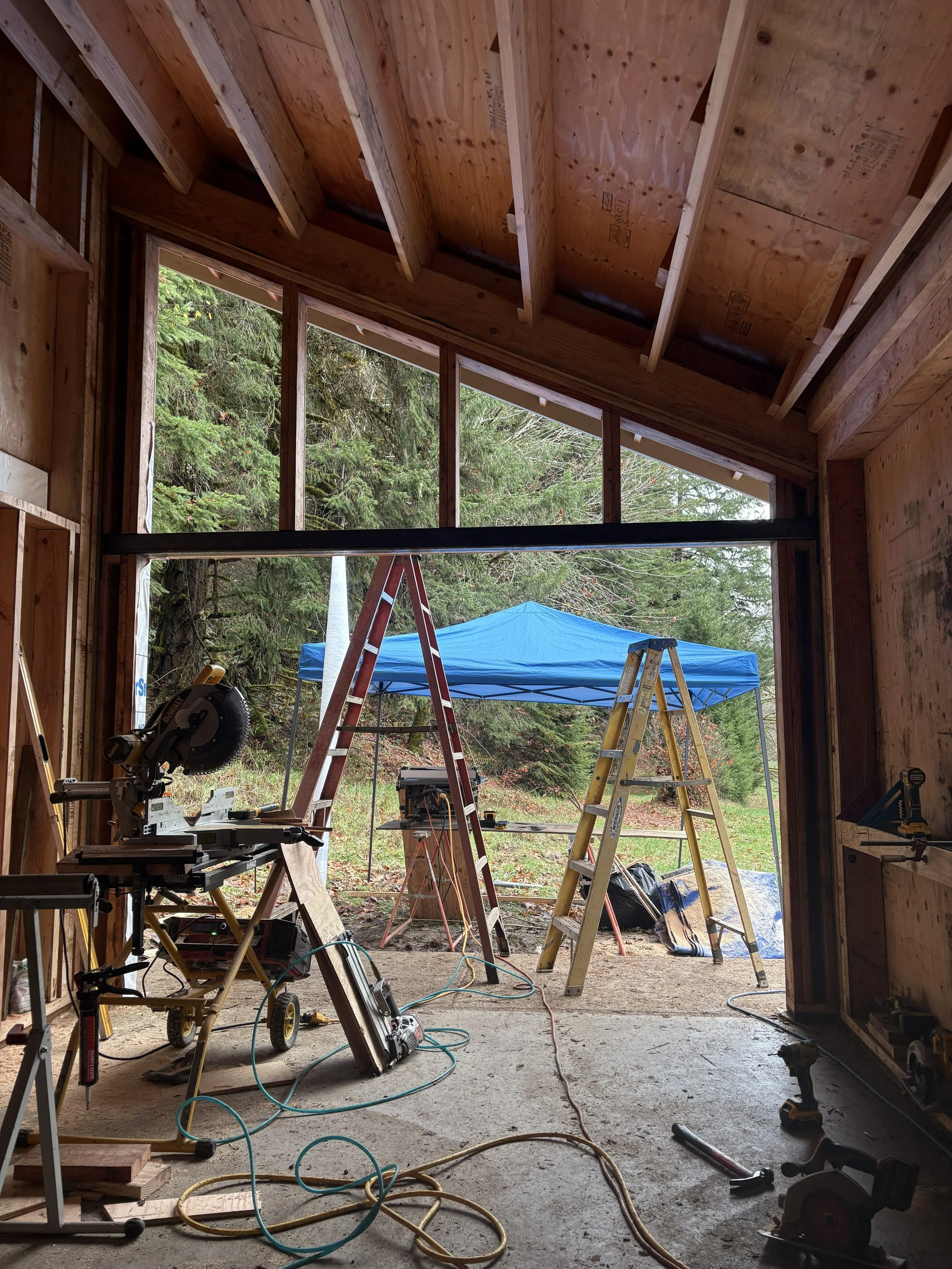 Construction site inside a building with wooden framing looks outside through open wall to trees with a blue tent, ladders, and construction tools and equipment scattered on the floor.