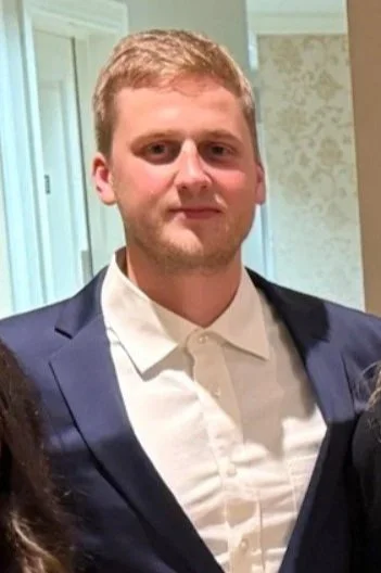 A young man with short, light-colored hair wearing a white dress shirt and a dark blazer, standing indoors.