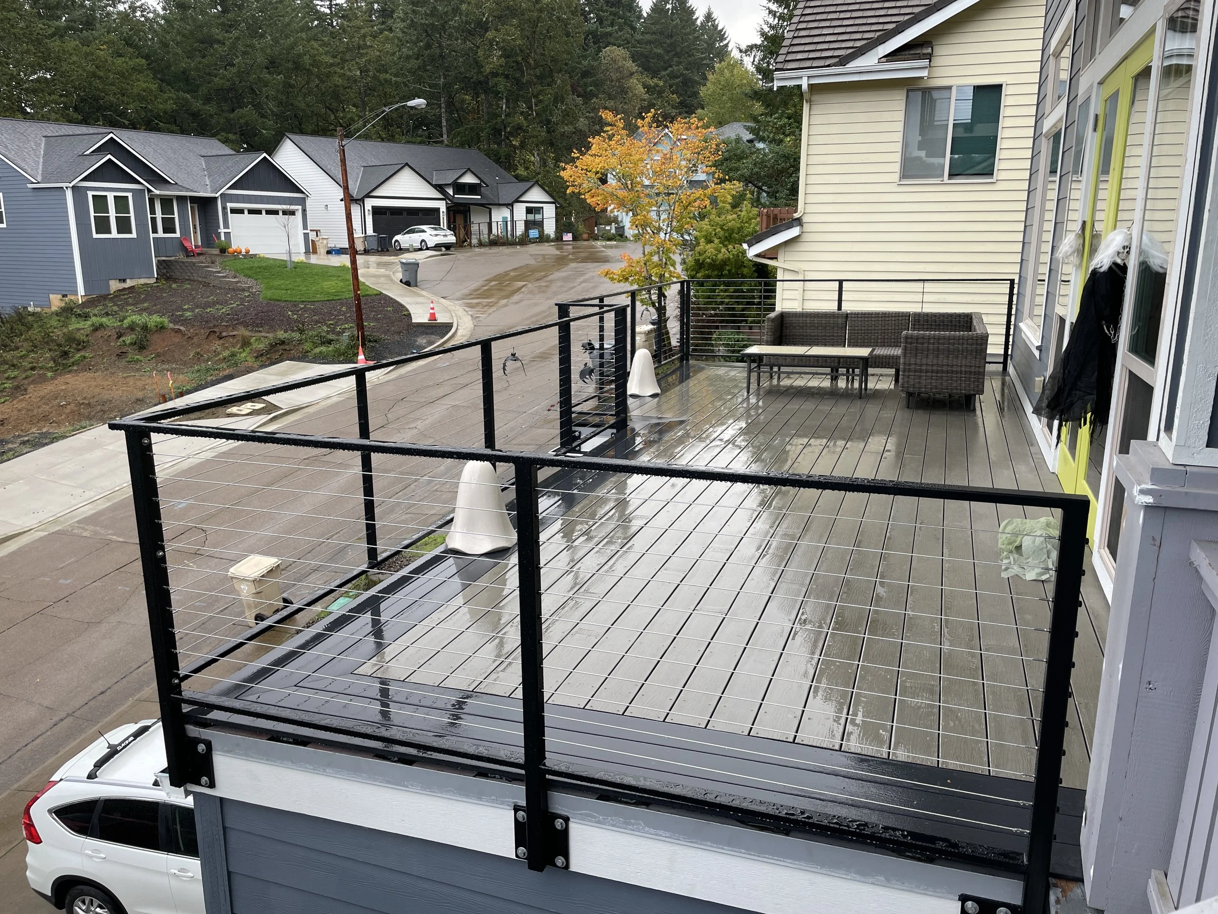 View from a balcony with a black railing overlooking a suburban street with houses, trees with autumn foliage, and a wet pavement after rain.