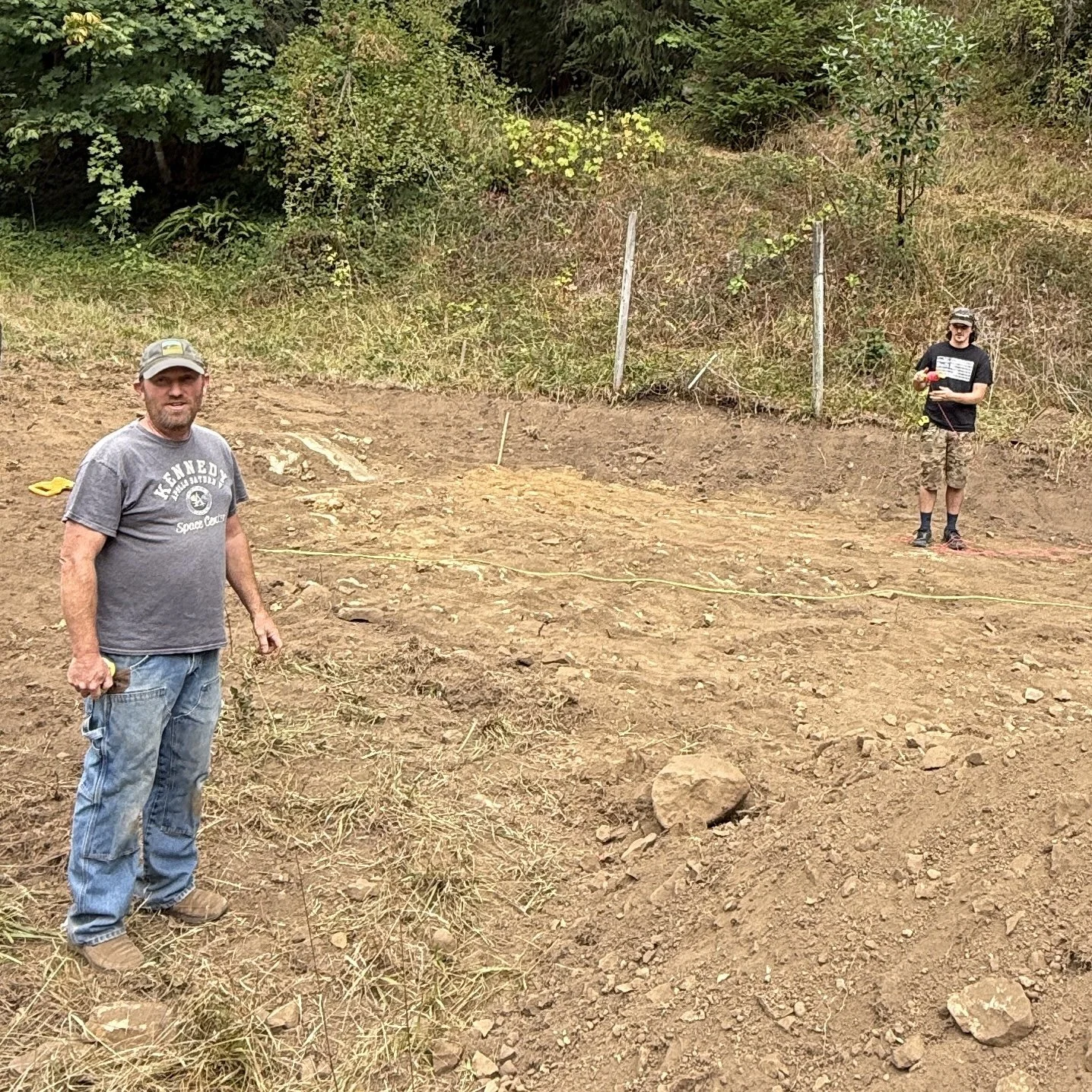 Two men standing in a cleared, rocky, dirt area with bushes and trees in the background. One man is closer to the camera, wearing a gray t-shirt and jeans, holding a small yellow object. The other man is farther away, wearing a black t-shirt and camo