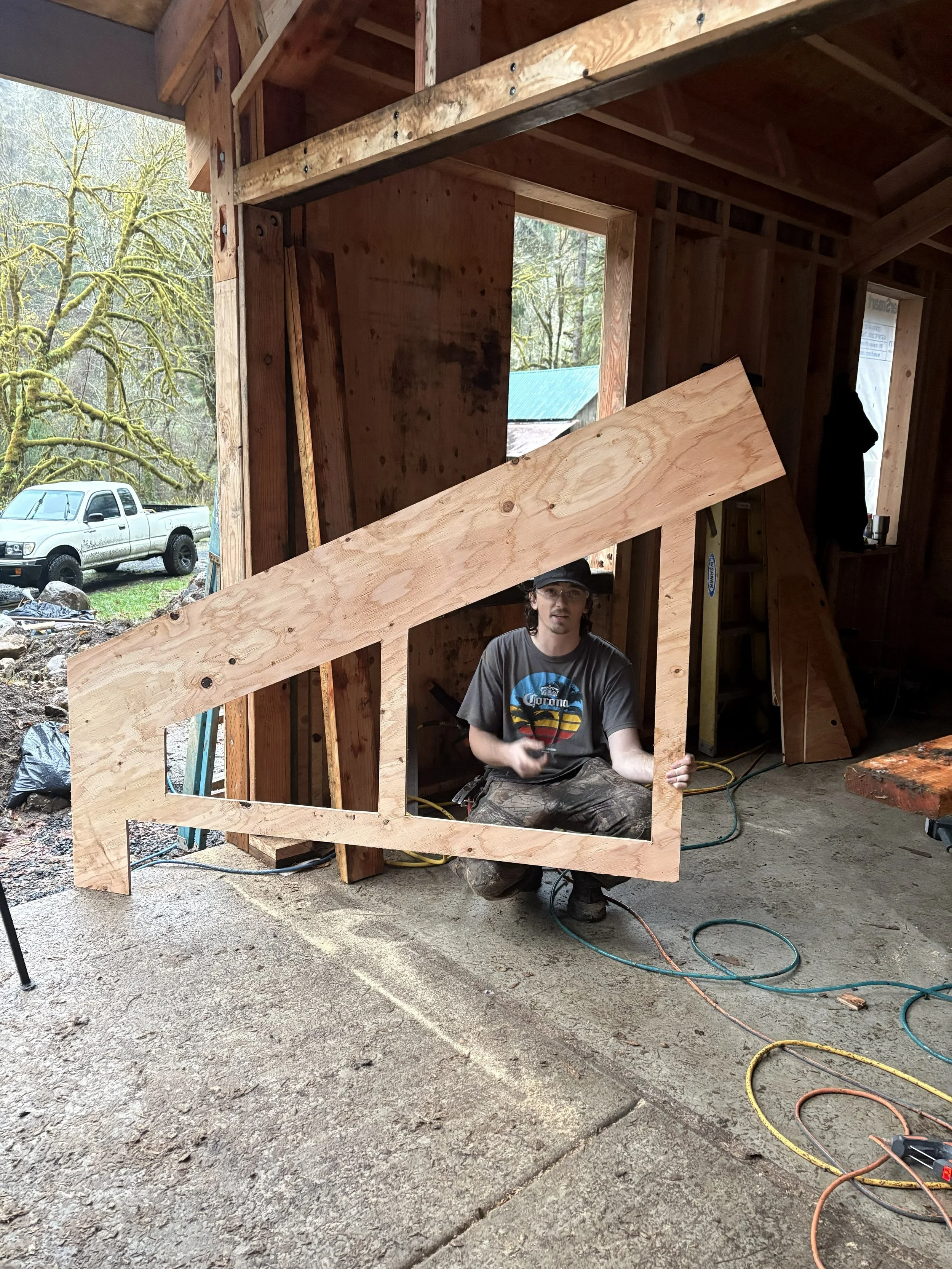 A man wearing a black cap and a dark gray t-shirt with a colorful logo, crouching in a partially constructed wooden frame of a building. The frame includes several large pieces of plywood and wooden beams. Outside, there is a snowy landscape with tre