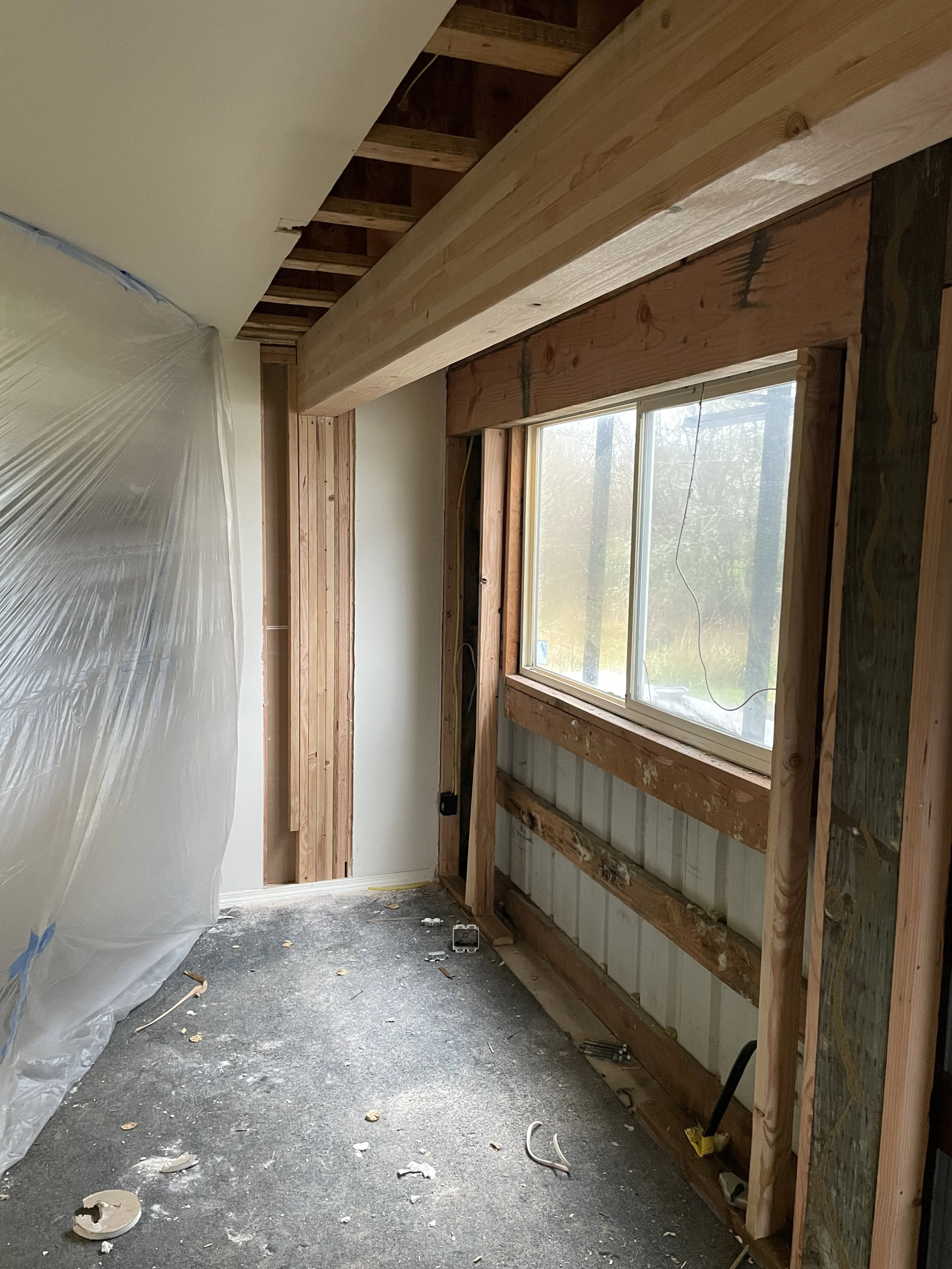 Interior of a room under renovation with exposed wooden framing around a window, partially covered wall, and construction debris on the floor. A large wooden beam installed against the ceiling.