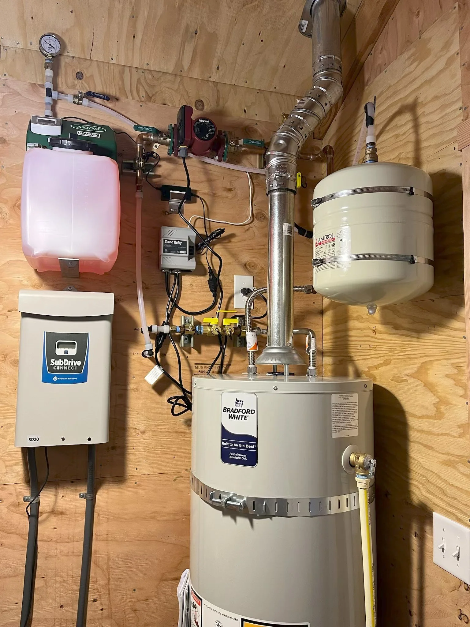 A water heater setup on a wooden wall with pipes, valves, a pressure gauge, and an electric control panel.
