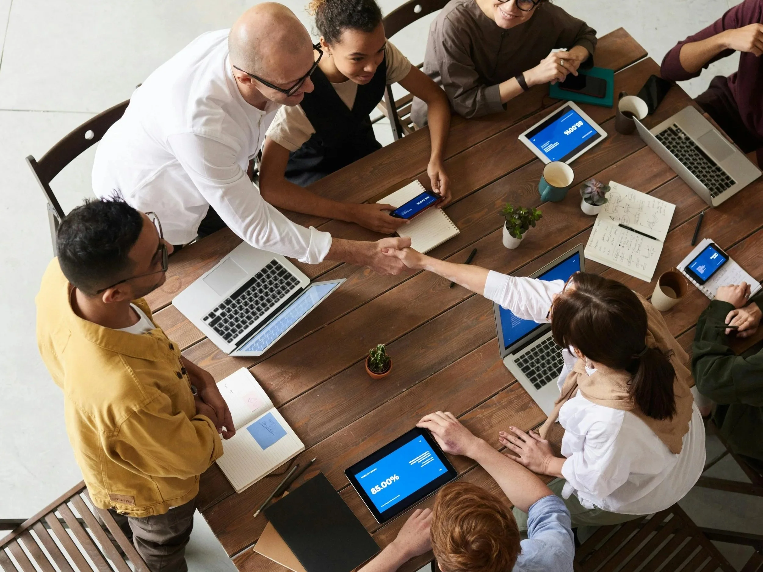 People at a meeting around a wooden table, shaking hands, with laptops, tablets, notebooks, and coffee cups.