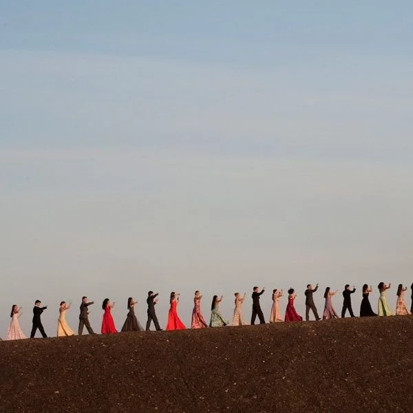 Grupo de personas caminando en fila por una colina, luciendo vestidos elegantes y trajes formales, en un día despejado.