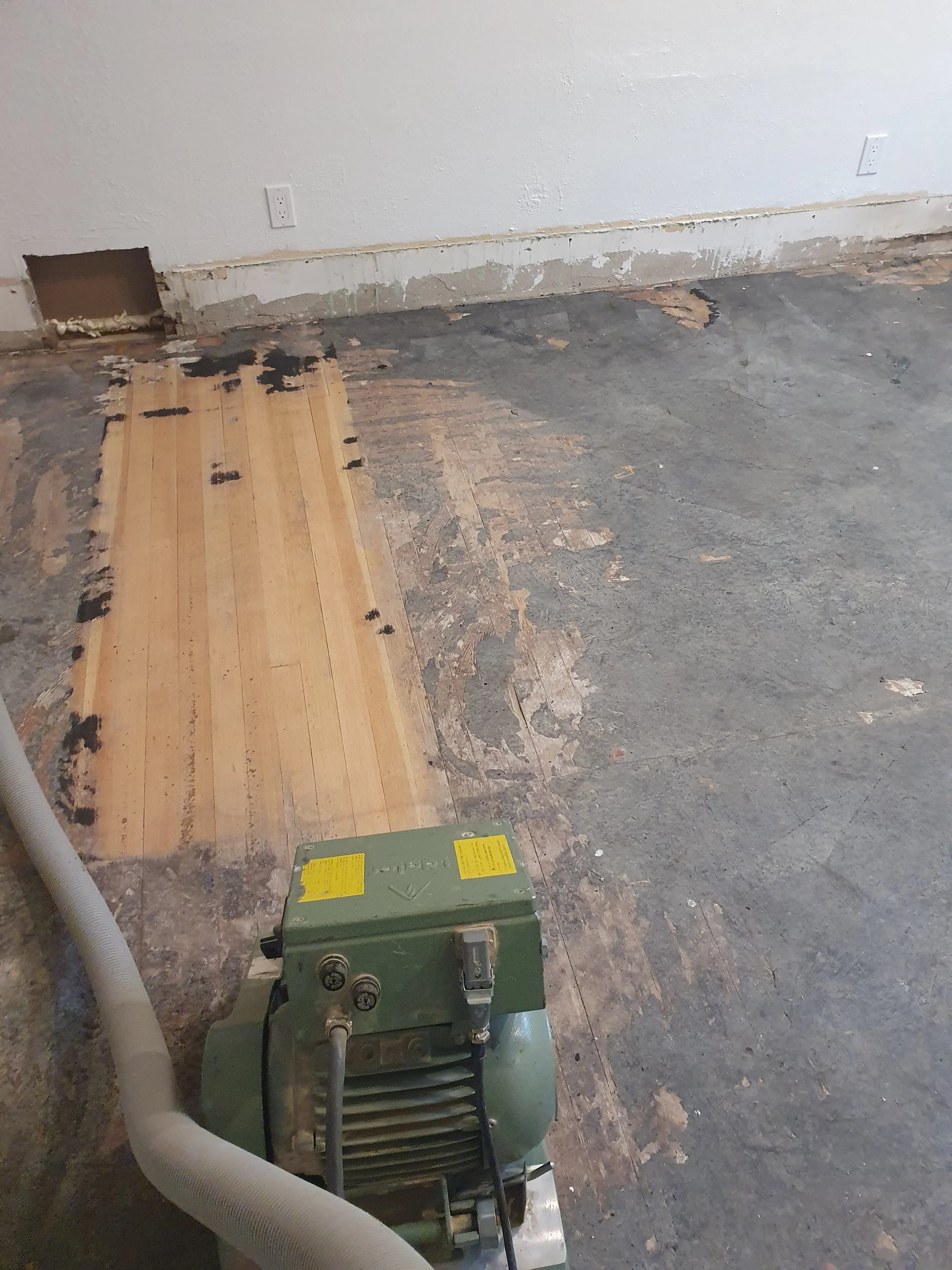 A room under renovation with exposed flooring, a partially removed old floor, and an area where new flooring is being installed. The image shows a green machine and hoses, along with electrical outlets on the wall.