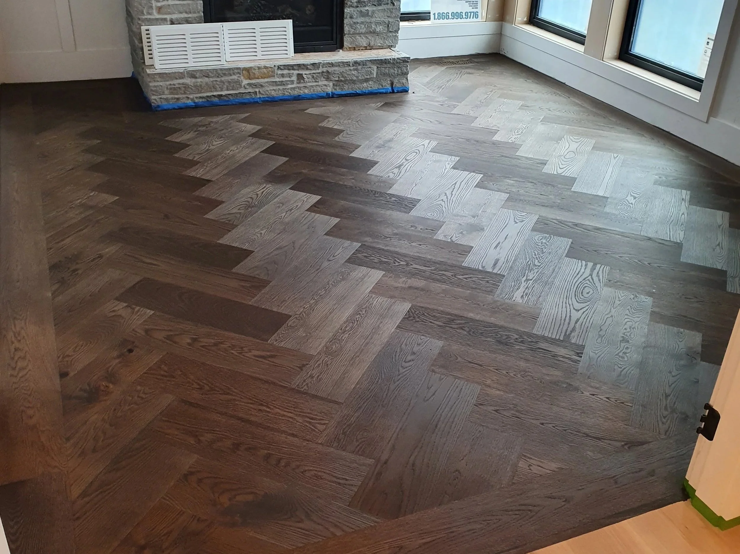 Interior of a room featuring a corner fireplace with a stone facade, large glass sliding doors, and a newly installed wood floor in a herringbone pattern.