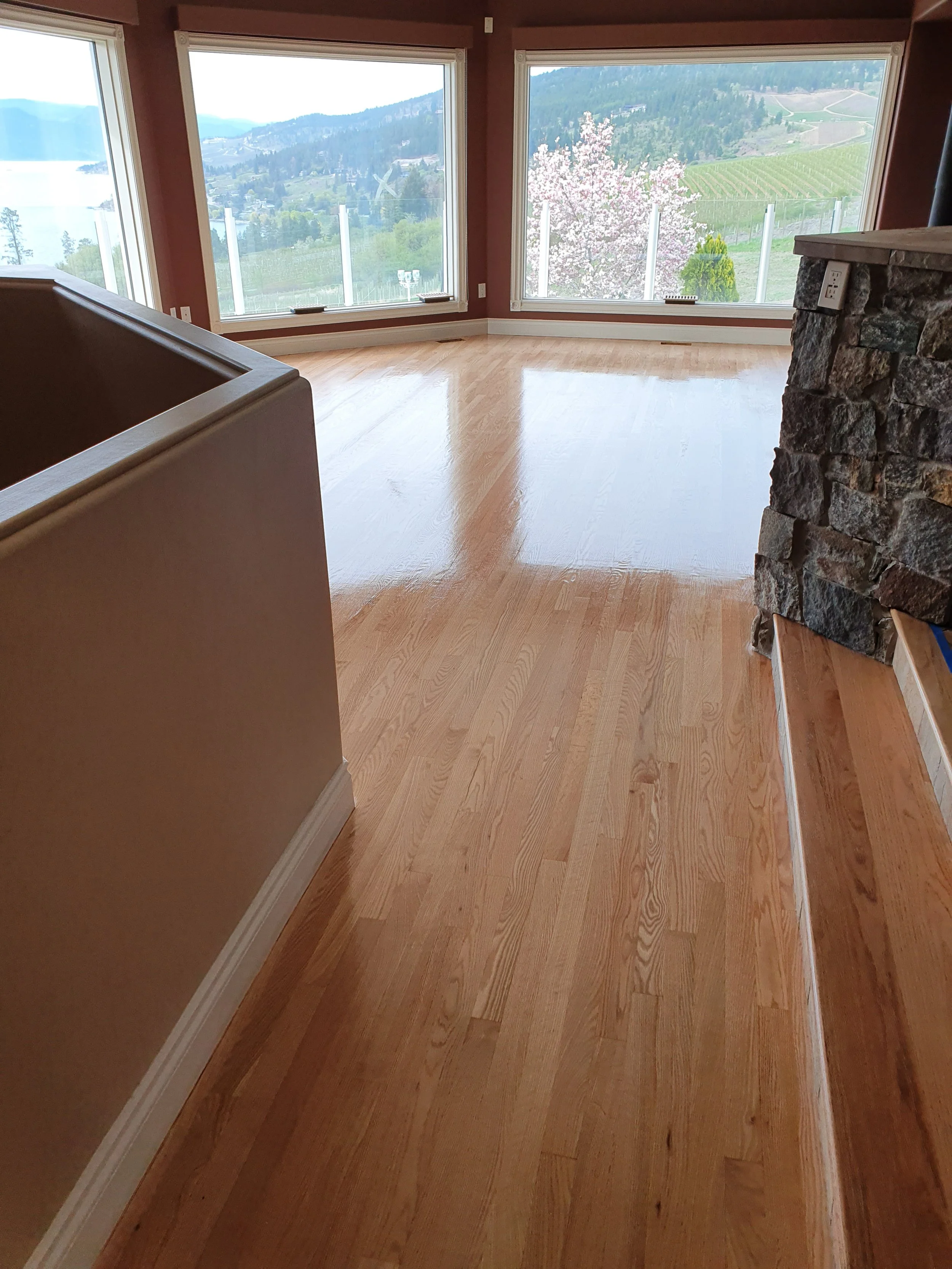 Empty living room with large windows overlooking a mountain landscape, hardwood floors, and a stone fireplace.