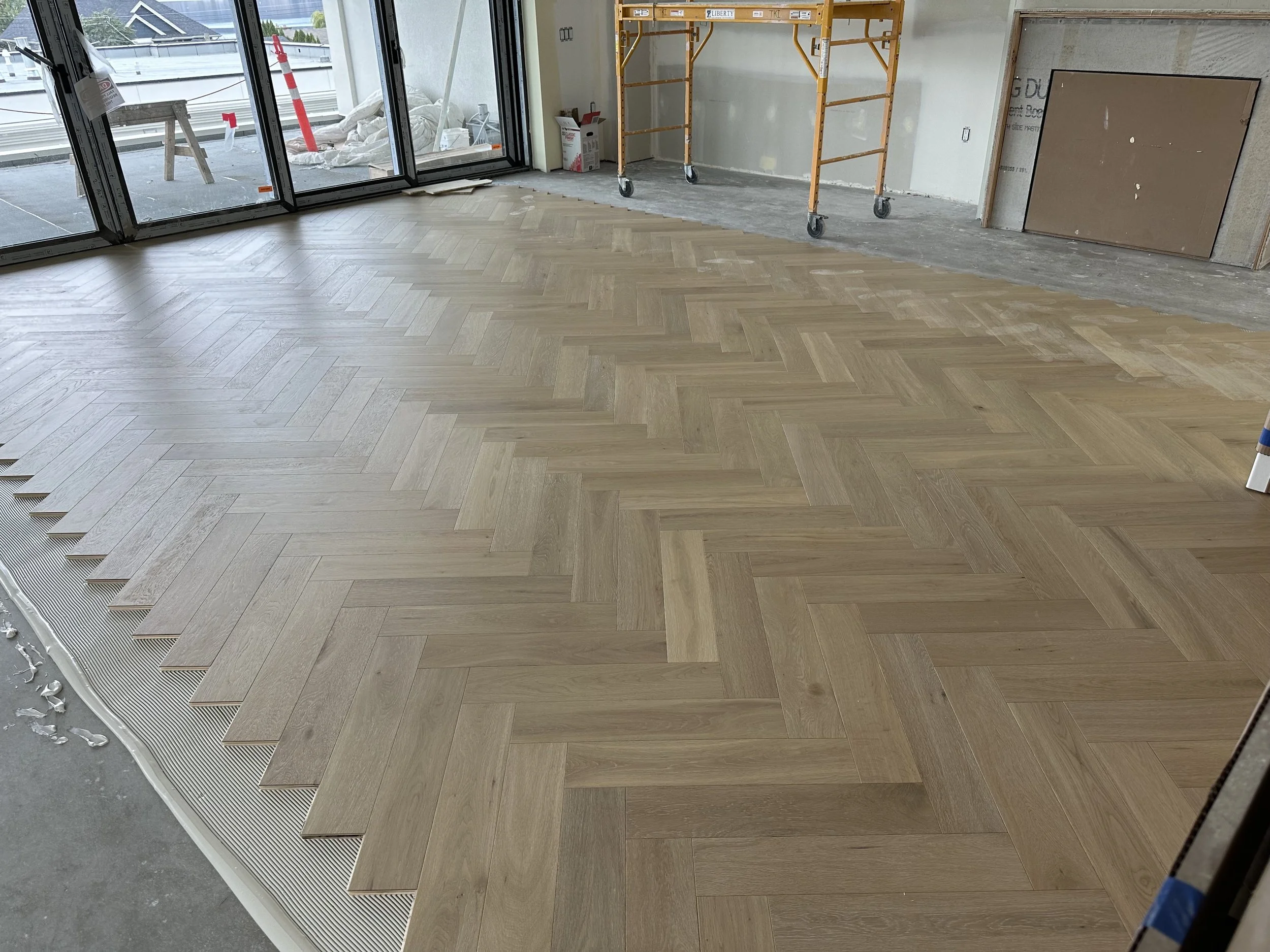 Interior of a room under construction with newly installed herringbone wood flooring and large windows, a scaffolding and construction materials visible.