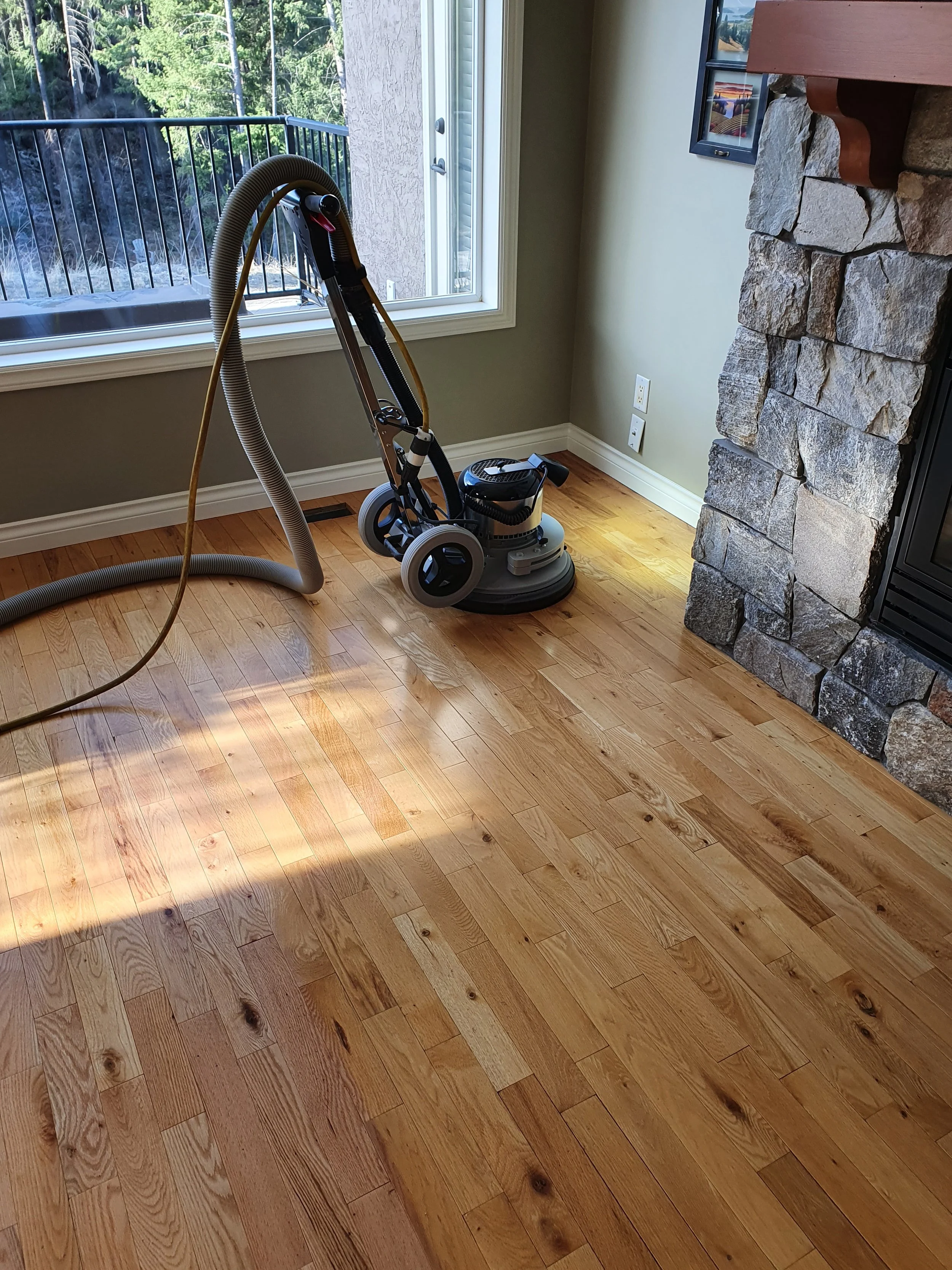 Empty living room corner with wooden floor, a wood and stone fireplace, a large window with a view of trees outside, and a vacuum cleaner in the corner.