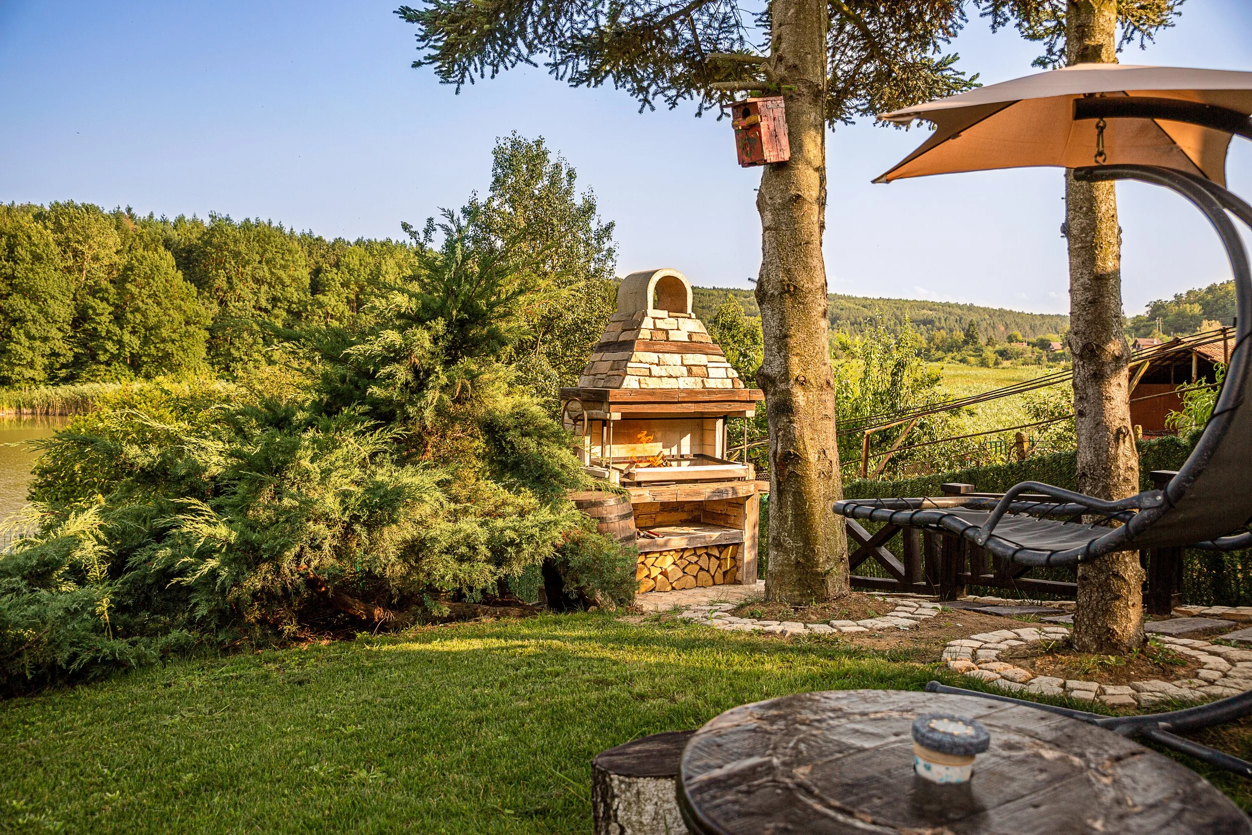 A backyard scene with a built-in brick outdoor pizza oven, surrounded by trees and lush greenery, with a nearby hammock and stone pathway.