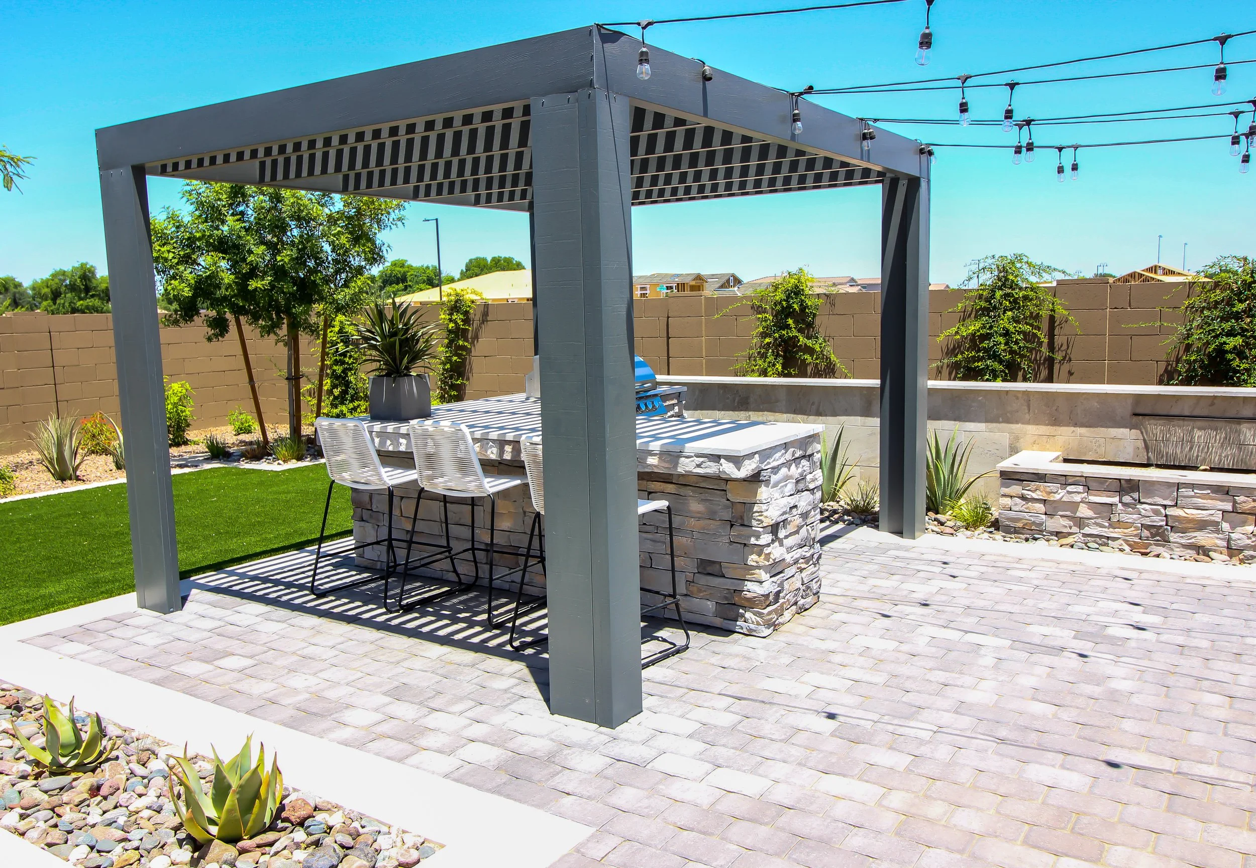Outdoor patio with a covered bar area featuring a stone counter, surrounded by patio chairs, string lights overhead, and landscaping with plants and a small lawn.